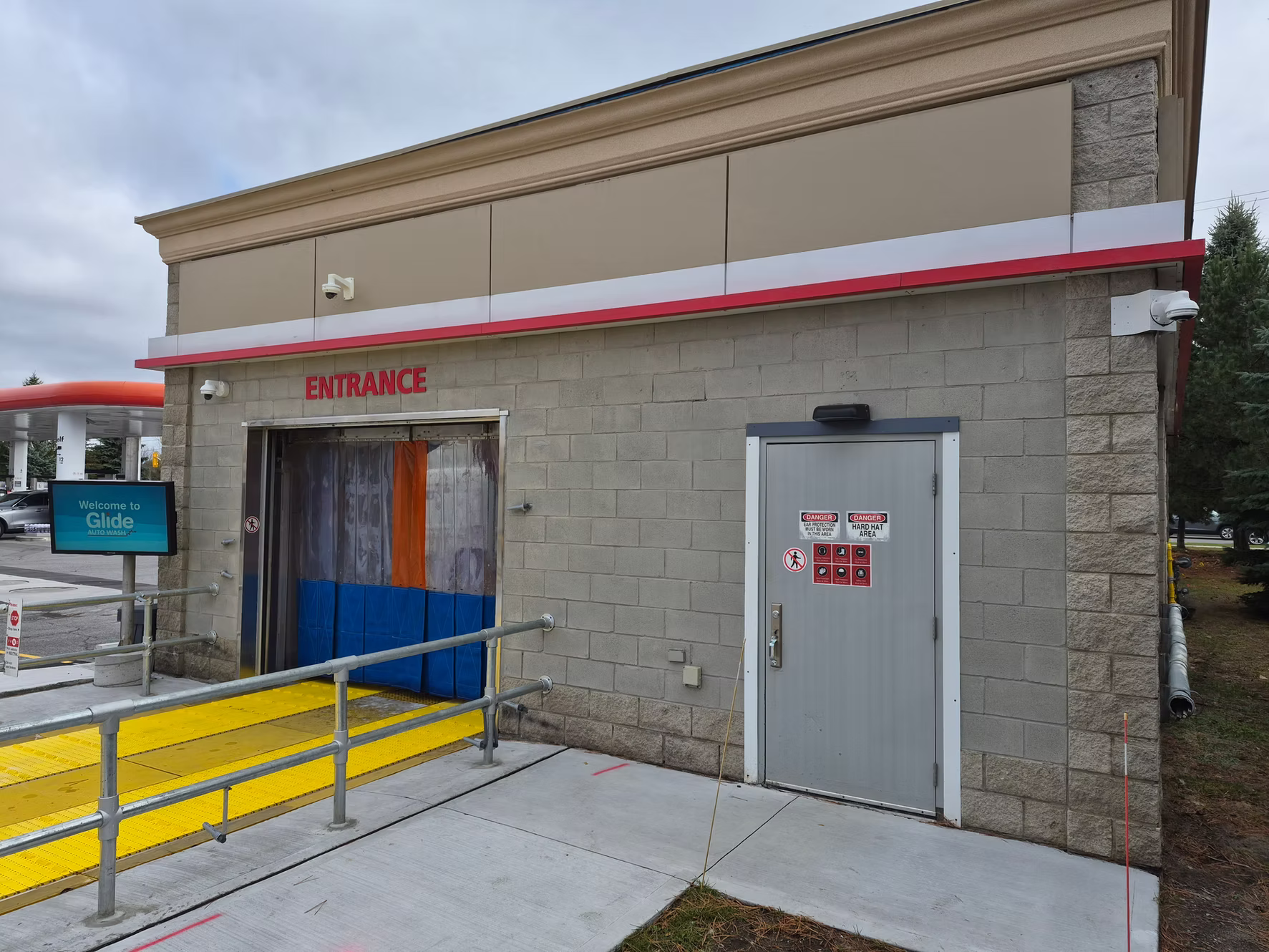 Exterior of a car wash facility with a sign that says 'Welcome to Glide Auto Wash'. There is an entrance with a yellow textured ramp and safety railings, and a gray door with warning signs. The building is made of brick with beige panels on top, and a security camera is mounted on the wall.