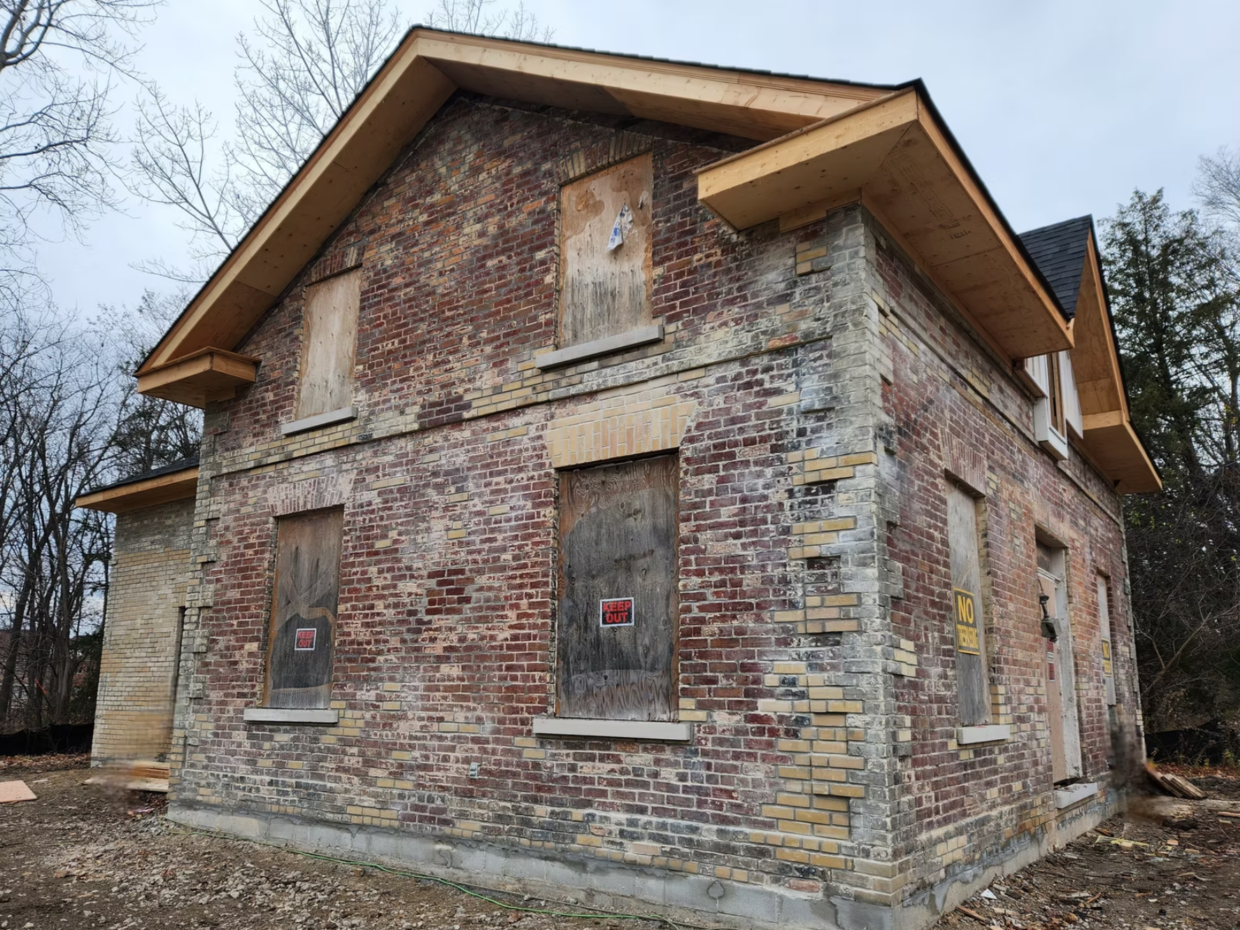 A two-story brick house under construction has boarded windows and doors, with signs reading 'KEEP OUT' and 'NO TRESPASSING.' The roof is partially finished with wood trim, and the sky is overcast. Leafless trees are nearby.