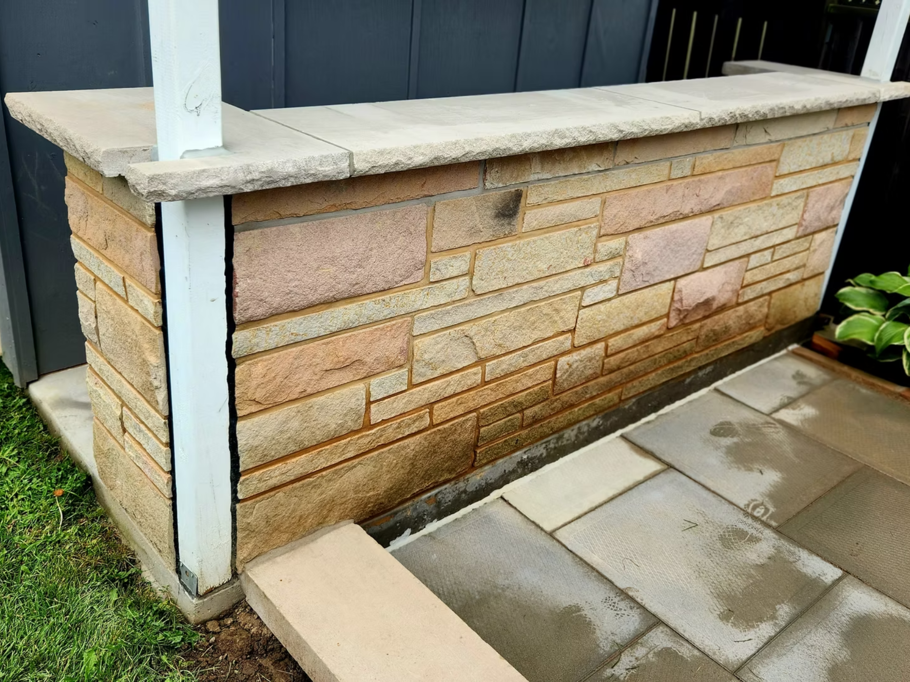 A brick and stone outdoor structure under construction, with paving stones laid in front and fencing in the background.