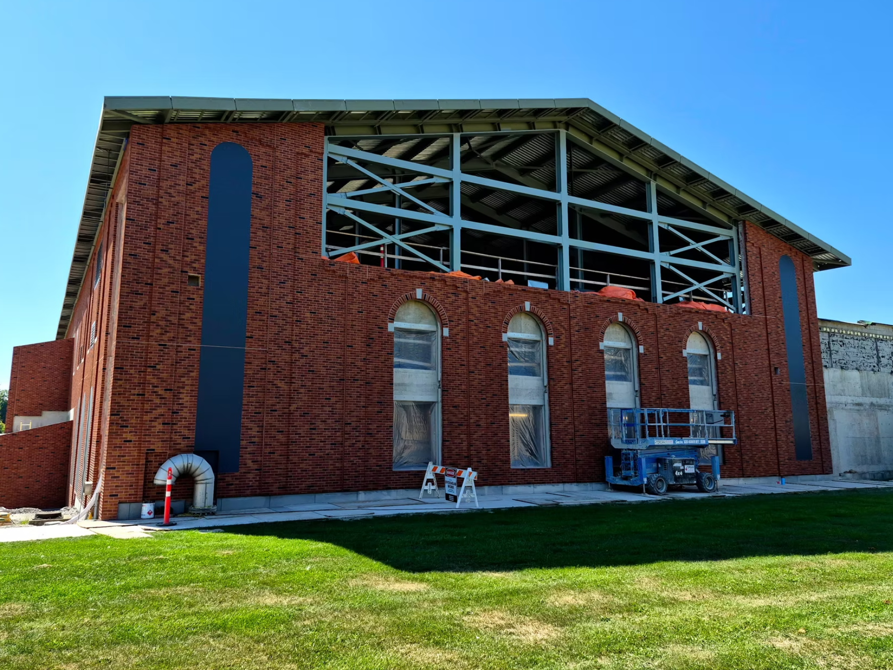 Building under construction with steel framework and brick exterior, a lift platform and construction barriers are present, with a grassy area in the foreground and a clear blue sky overhead.