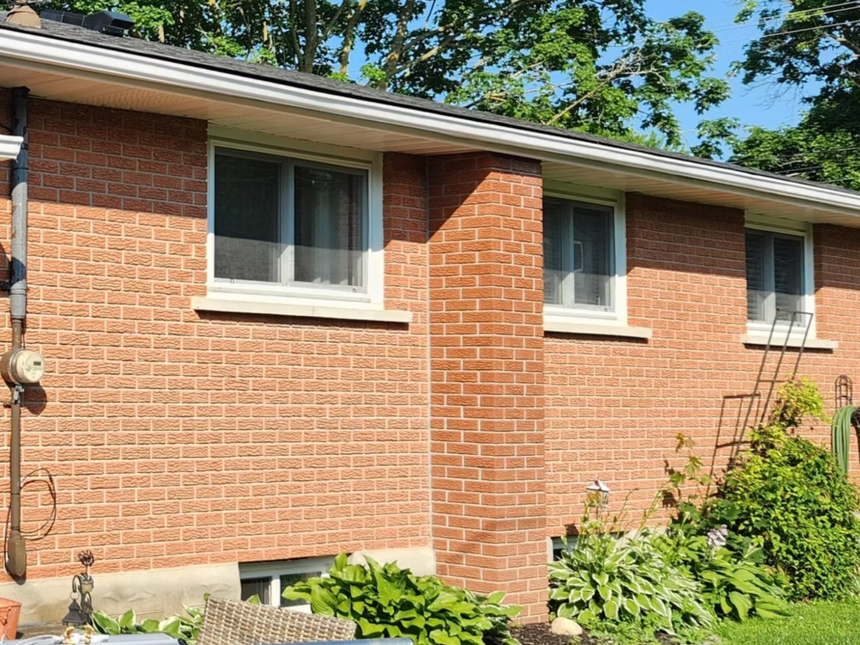 The image shows the exterior of a brick house with two windows, a gutter, and a ladder leaning against the wall. There are green trees and bushes in the yard, with a garden hose and outdoor furniture visible.