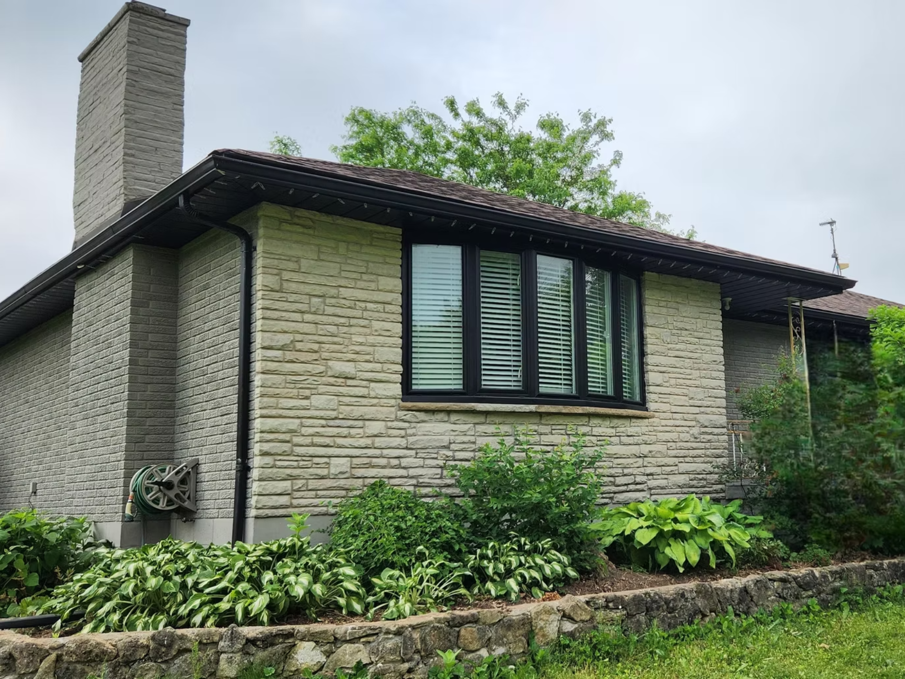 The side of a house with a stone exterior, a window with black trim and blinds, a chimney, and a garden with green plants and shrubs.