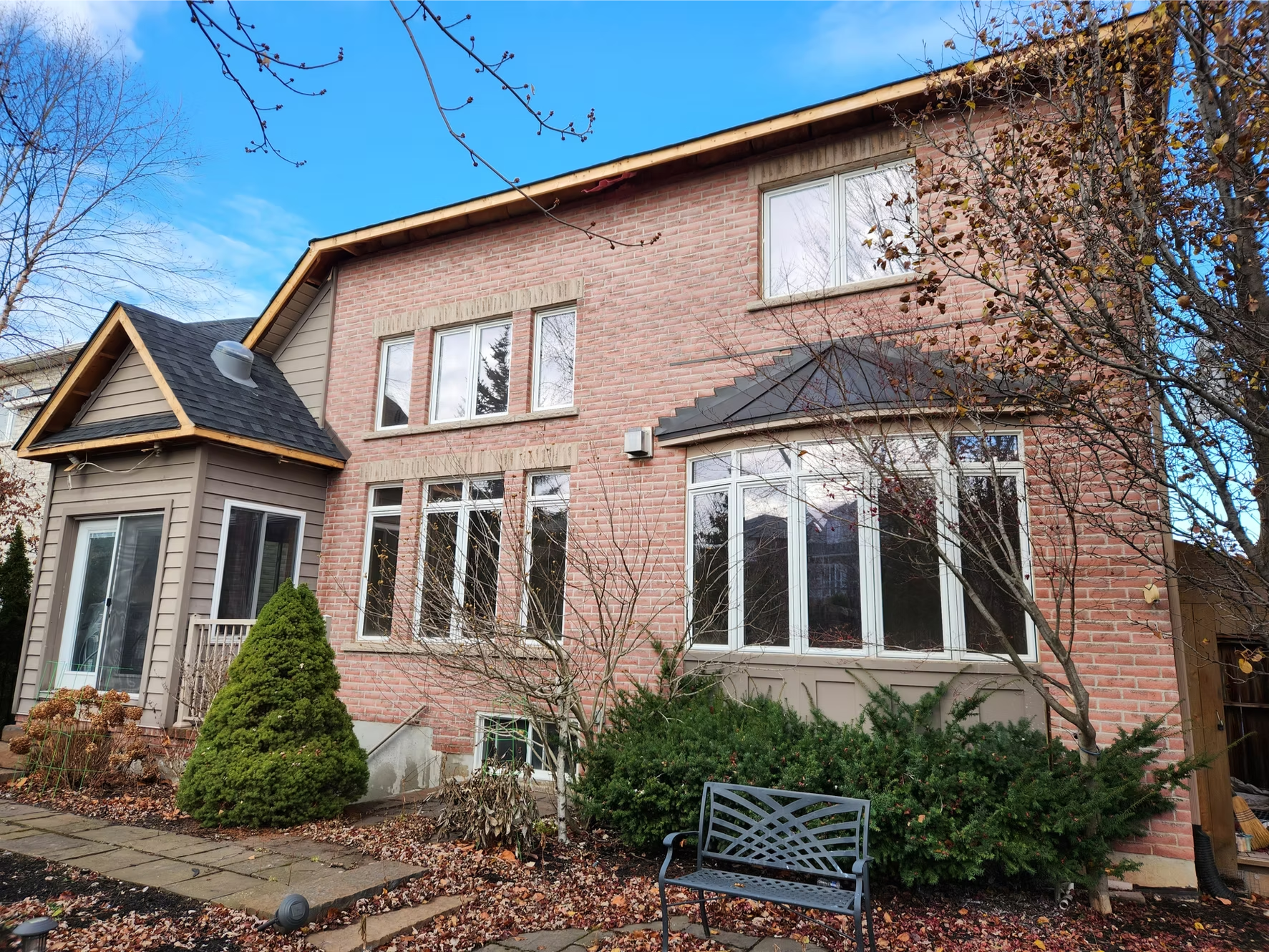 A two-story house with a brick exterior, large windows, and a small front yard with trees, bushes, a black metal bench, and a stone pathway.