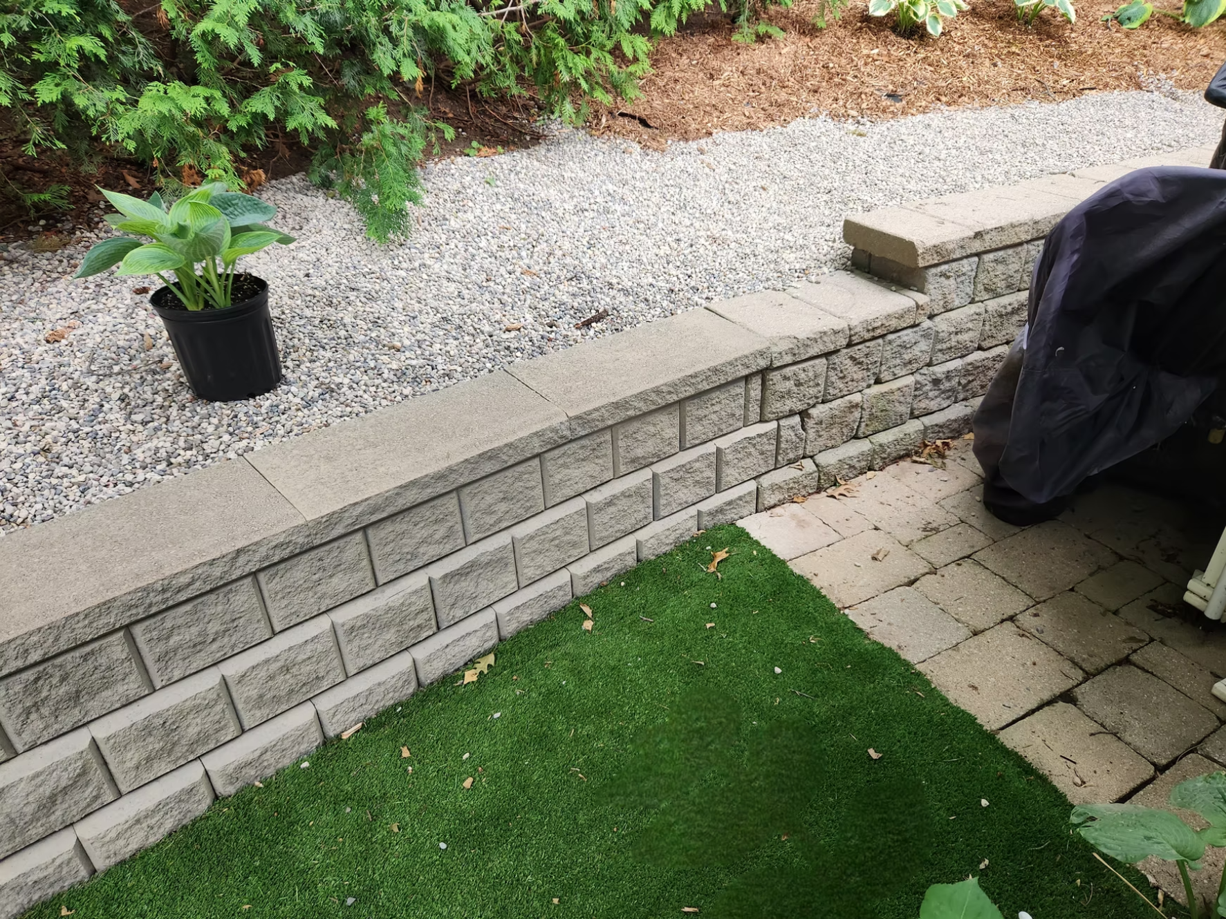 A landscaped backyard with a low brick wall, gravel bed, potted plant, artificial grass patch, and paver patio area.