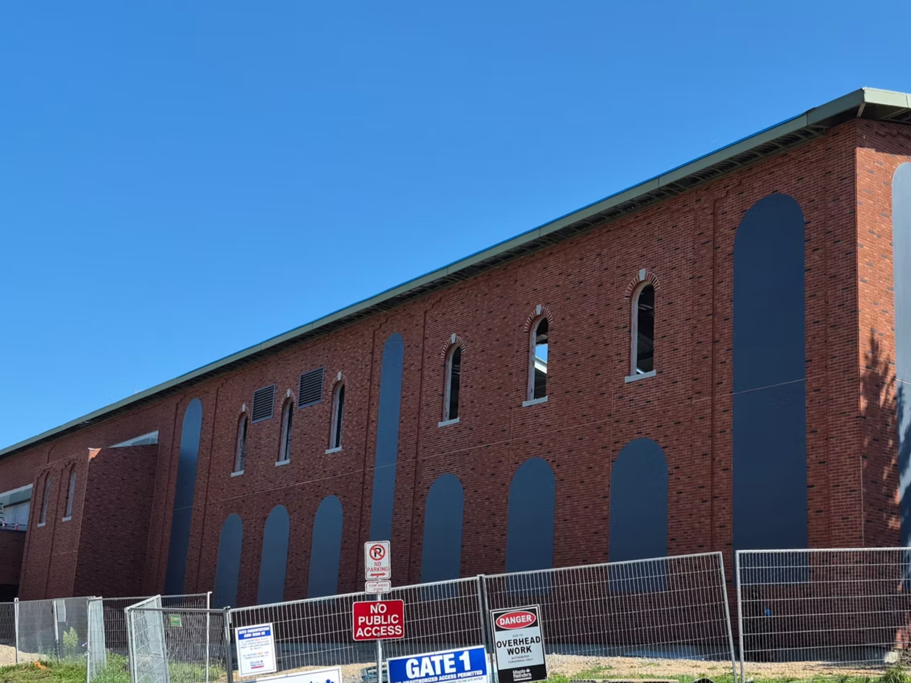 A large red brick building with arched windows and grey accents, surrounded by a construction fence with multiple signs that read 'No Public Access' and 'Danger Overhead Work'. The sky above is clear and blue.