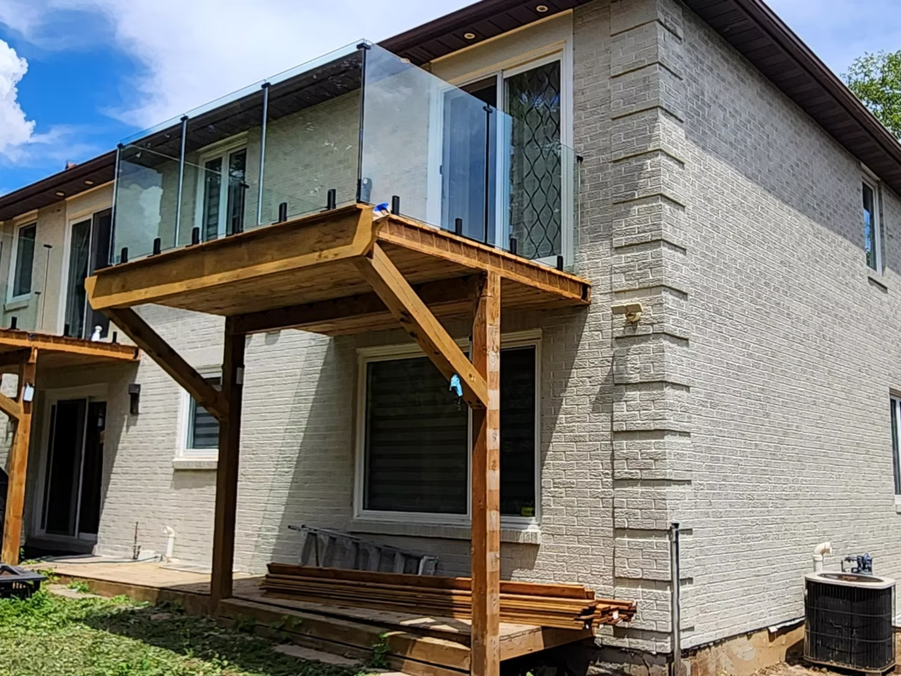 A brick house with a balcony under construction. The balcony has glass panels and wooden supports. There are construction materials and tools around, including wooden planks and a ladder.