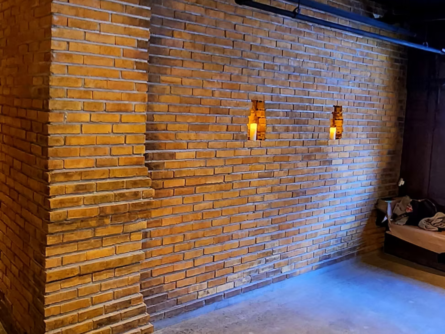 Interior brick wall with two small rectangular cutouts containing candles, a bench with clothes and bag on it, and wood beams on the ceiling.