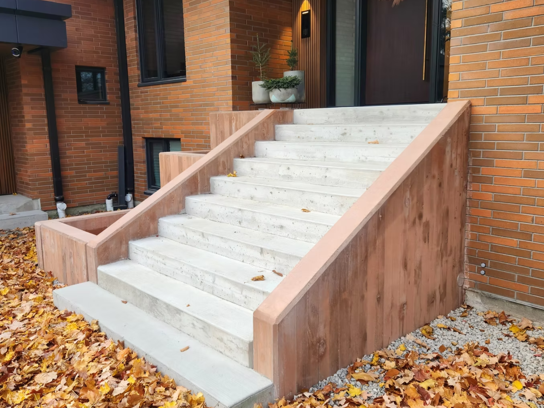 Concrete stairs with wooden sides leading up to a modern house entrance, decorated with potted plants, and surrounded by fallen autumn leaves.