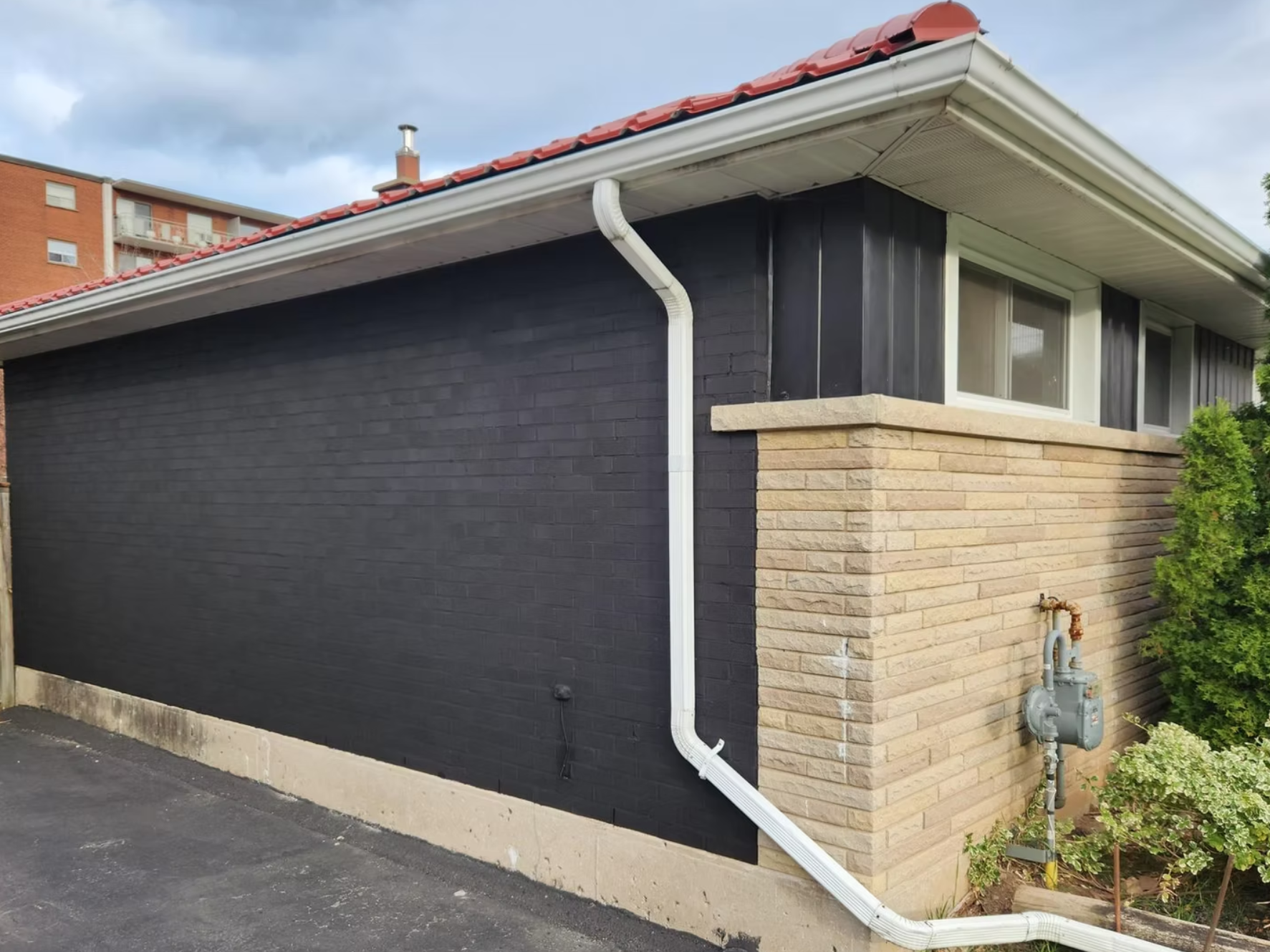The exterior of a house with black painted brick wall and beige brick corner, a white gutter system, and a gas meter with pipes. There are windows with white frames and a small green shrub nearby.