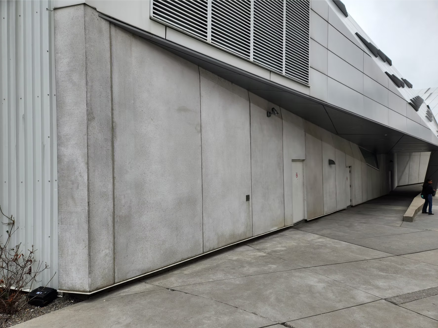 A modern building with concrete walls and metal ventilation panels, featuring security cameras and a person walking nearby.