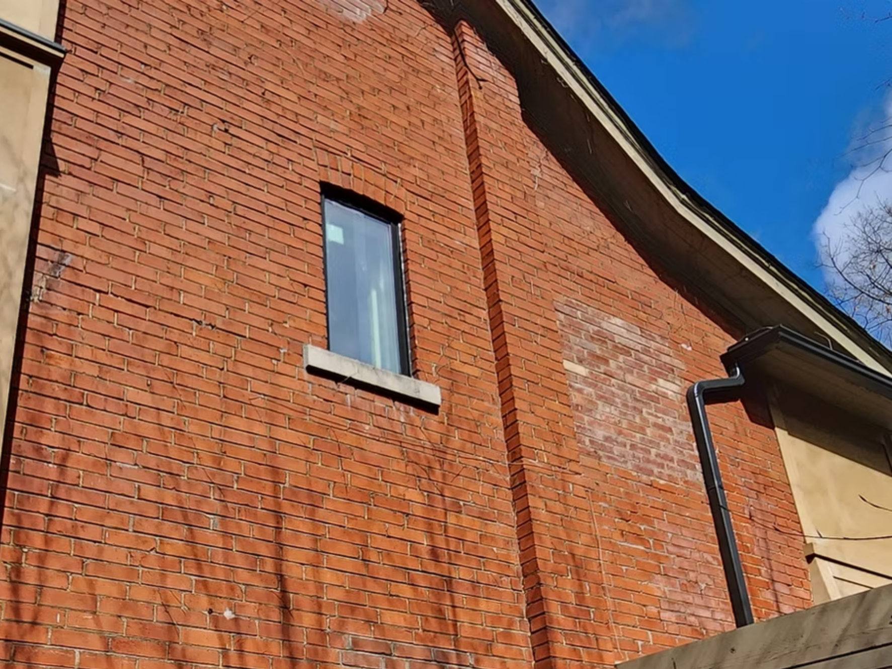 Close-up of a brick house wall with a window and part of a roof, under a blue sky with some clouds.