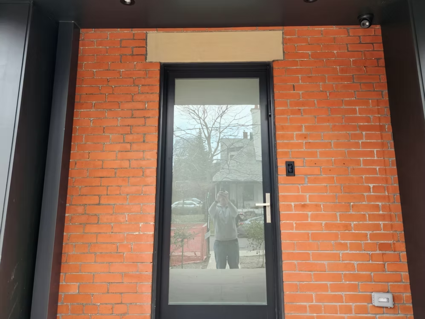 A door with a glass window set into a red brick wall. The reflection in the glass shows a person taking the photo, trees, and a residential street scene.