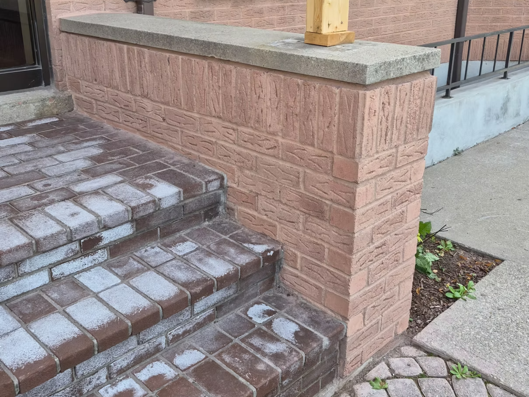 Brick steps leading up to a building entrance, with a brick and stone wall, a concrete ledge, and a wooden post.