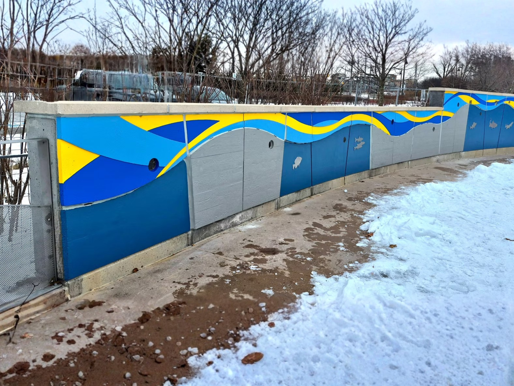 A colorful outdoor wall mural featuring blue and yellow wavy patterns with fish silhouettes, located along a concrete barrier near a snow-covered sidewalk and leafless trees in the background.