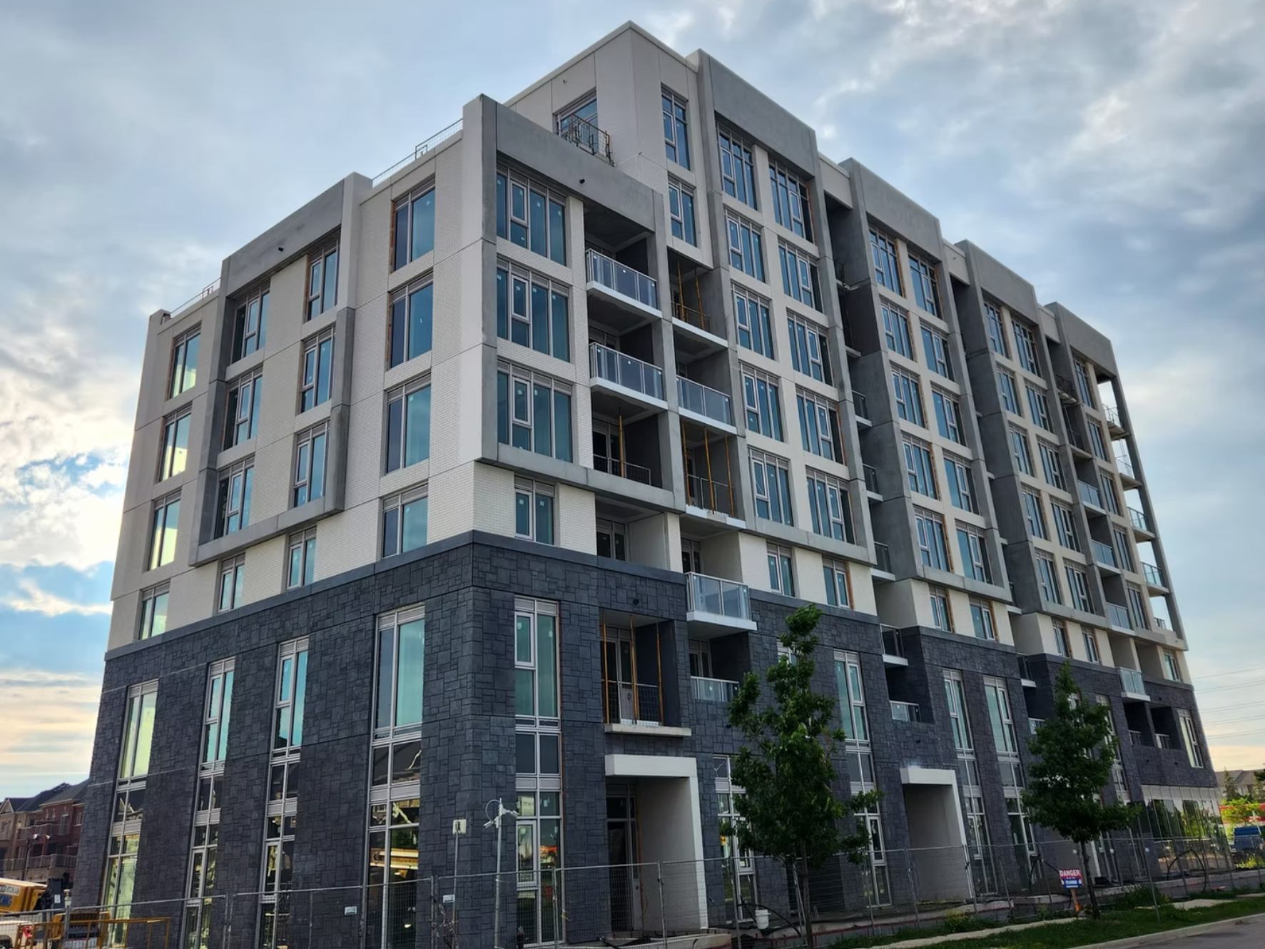 A multi-story modern apartment building with large windows and balconies, under a partly cloudy sky with some trees in the foreground.