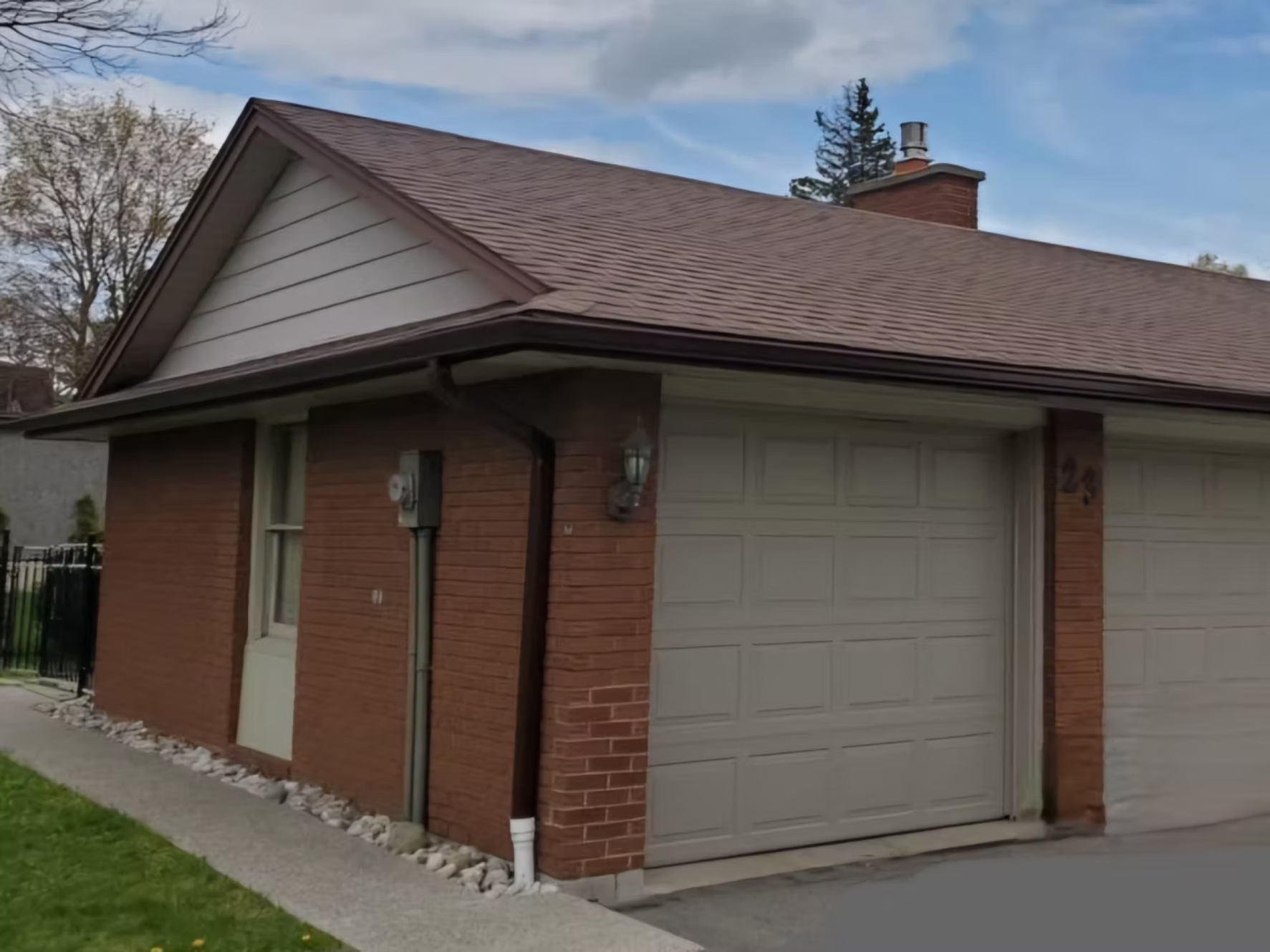 A house with a brick exterior and an attached two-car garage with a white door. The roof is brown with shingles, and there is a brick chimney. There are two windows on the house and a light fixture beside the garage door. The house has a small strip of rock landscaping along the edge and a sidewalk in front. There are trees in the background and a partly cloudy sky.