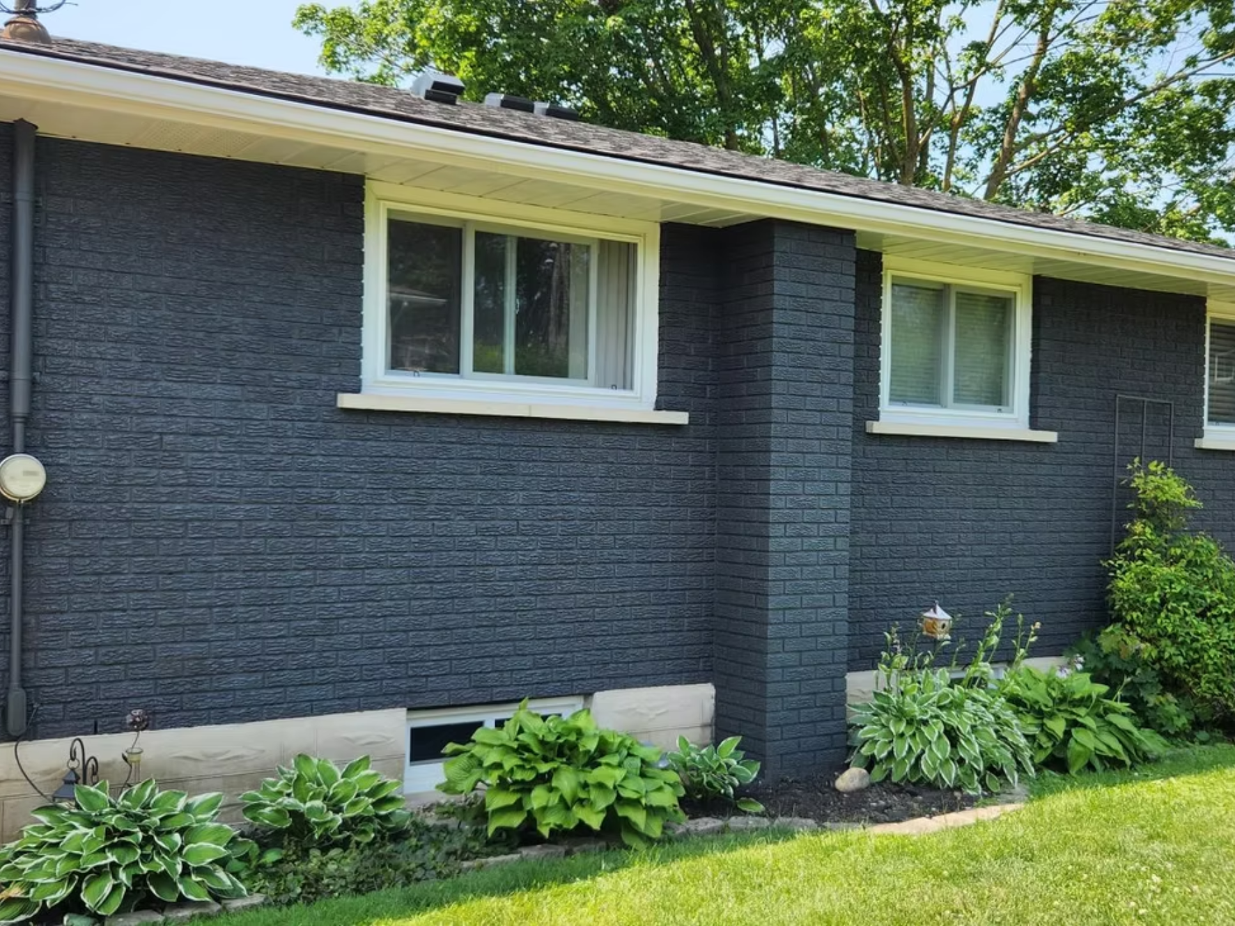 The side of a house painted dark gray with two rectangular windows. The house has a roof with shingles, and there are green trees in the background. There are plants and bushes in the garden in front of the house with green grass.
