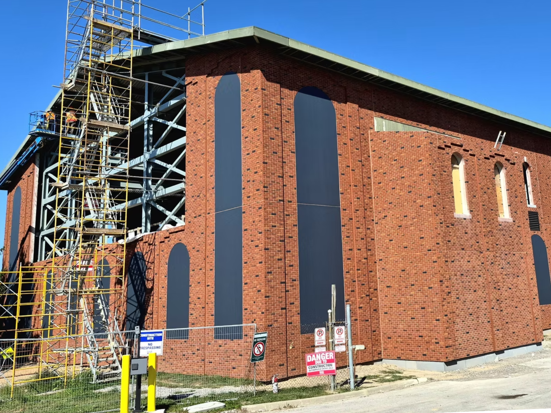A brick building under construction with scaffolding on the left side. The building has arched windows covered with black material. Construction signs and safety barriers are at the front, including warnings and restrictions.