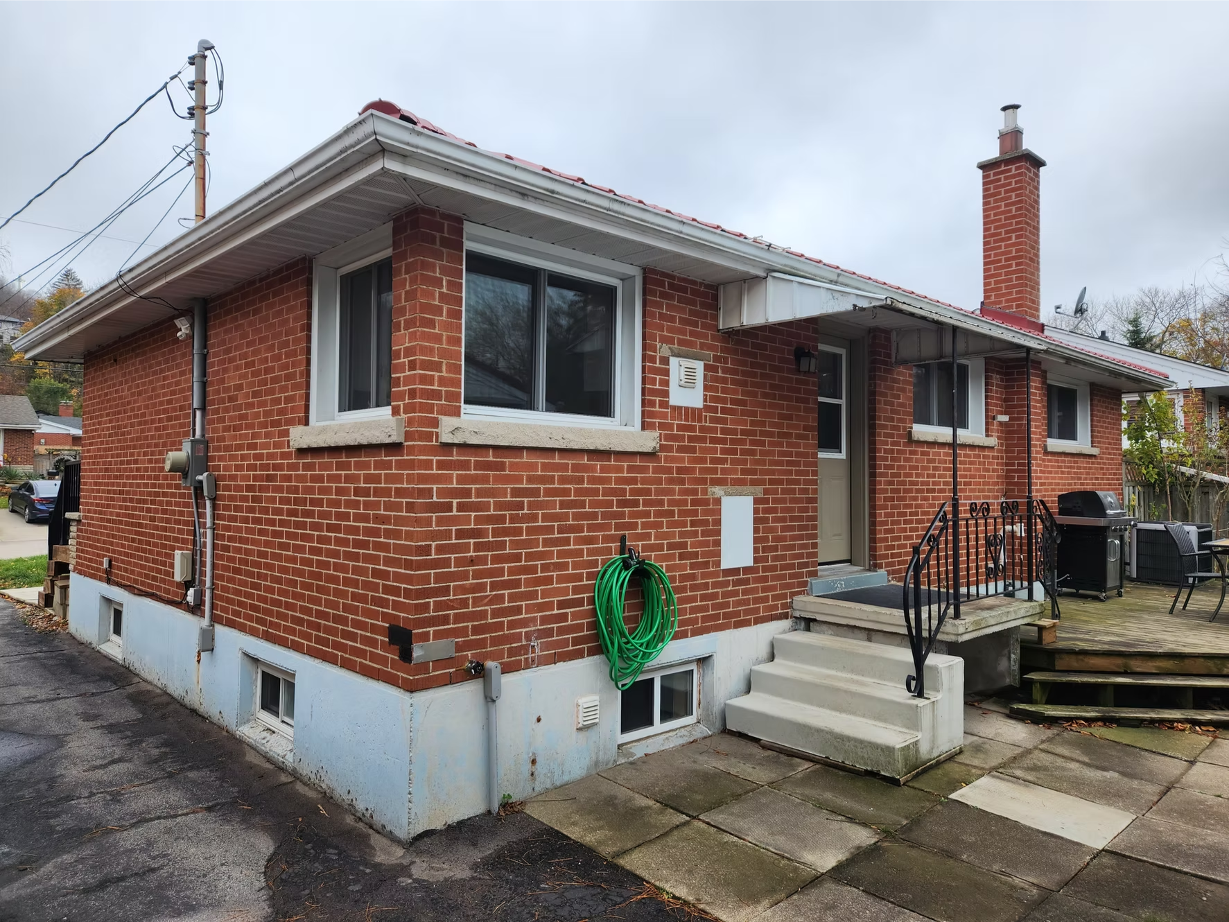 Red brick house with small steps leading to entrance, black metal railing, satellite dish, green garden hose, black grill, and a deck area, with overcast sky in the background.