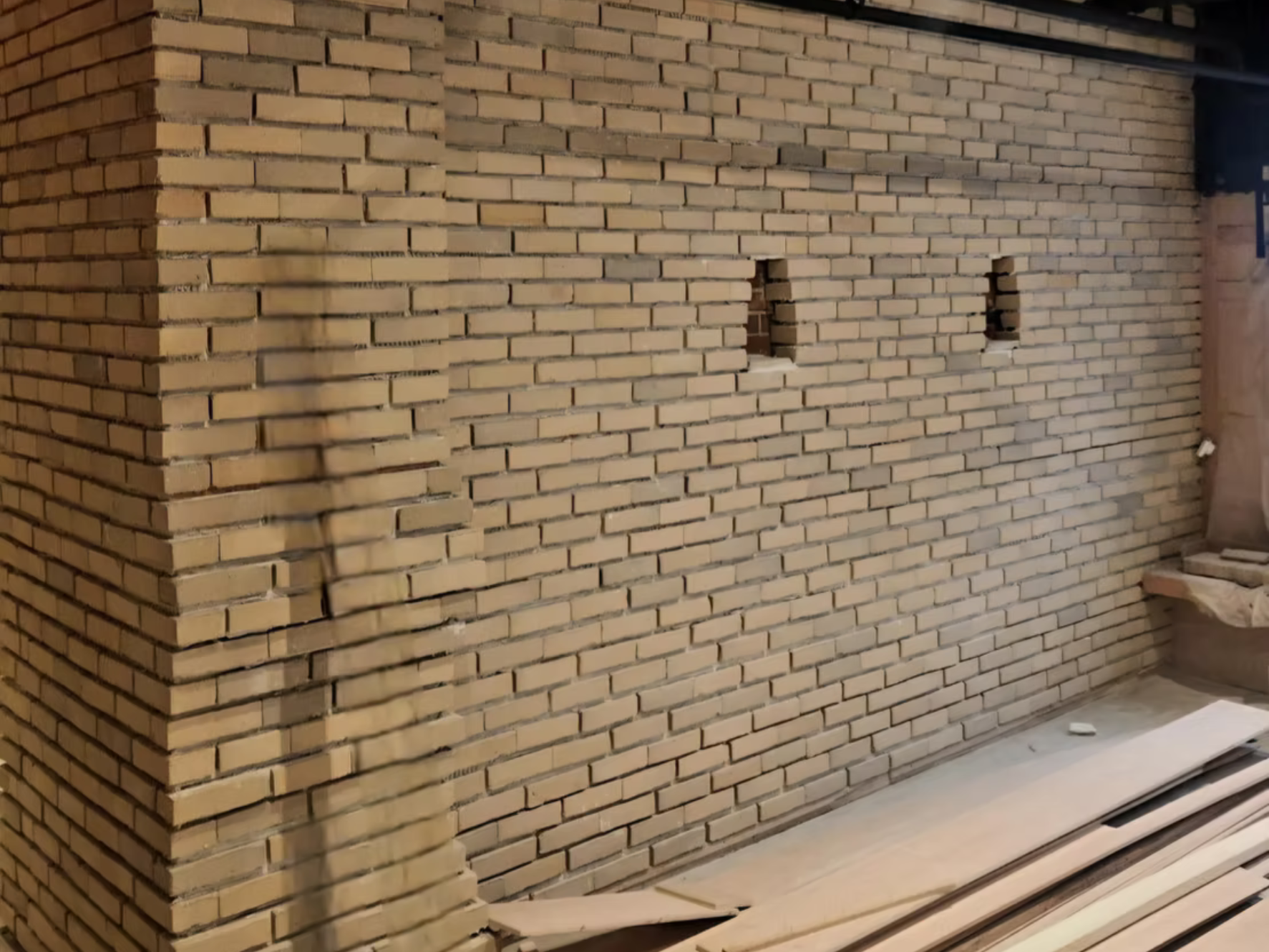 Interior brick wall with two small rectangular openings, construction materials and tools on the floor.