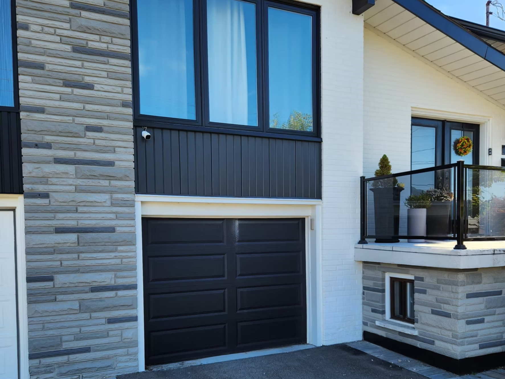 Front view of a modern house featuring a black garage door, large windows, a balcony with potted plants, white brick and stone siding, and a decorated wreath on the door.