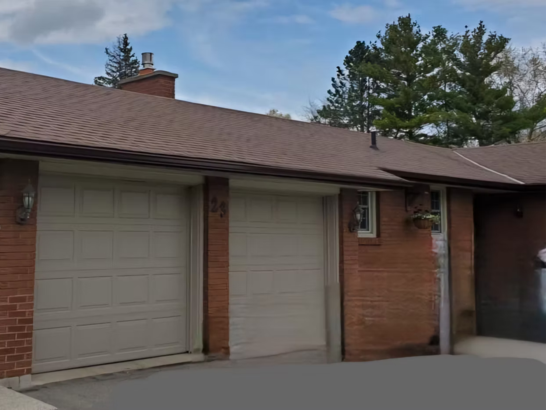 A brick house with a beige garage door, a small window with flower box, and house number 24. The house has a brown shingle roof and is surrounded by tall trees, with a partly cloudy sky overhead.