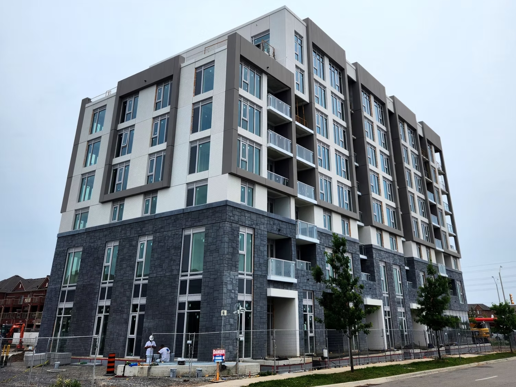 Newly constructed modern multi-story residential building with glass balconies, dark stone base, and gray upper floors under construction, with workers and a fenced area in front.