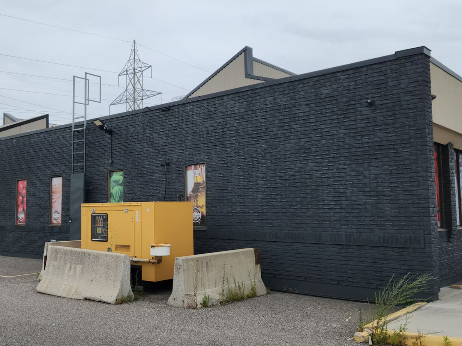 A black brick building with small rectangular windows displaying colorful graphics; in front, there are concrete barriers, a yellow utility box, and an electrical meter. In the background, there are power lines and a cloudy sky.