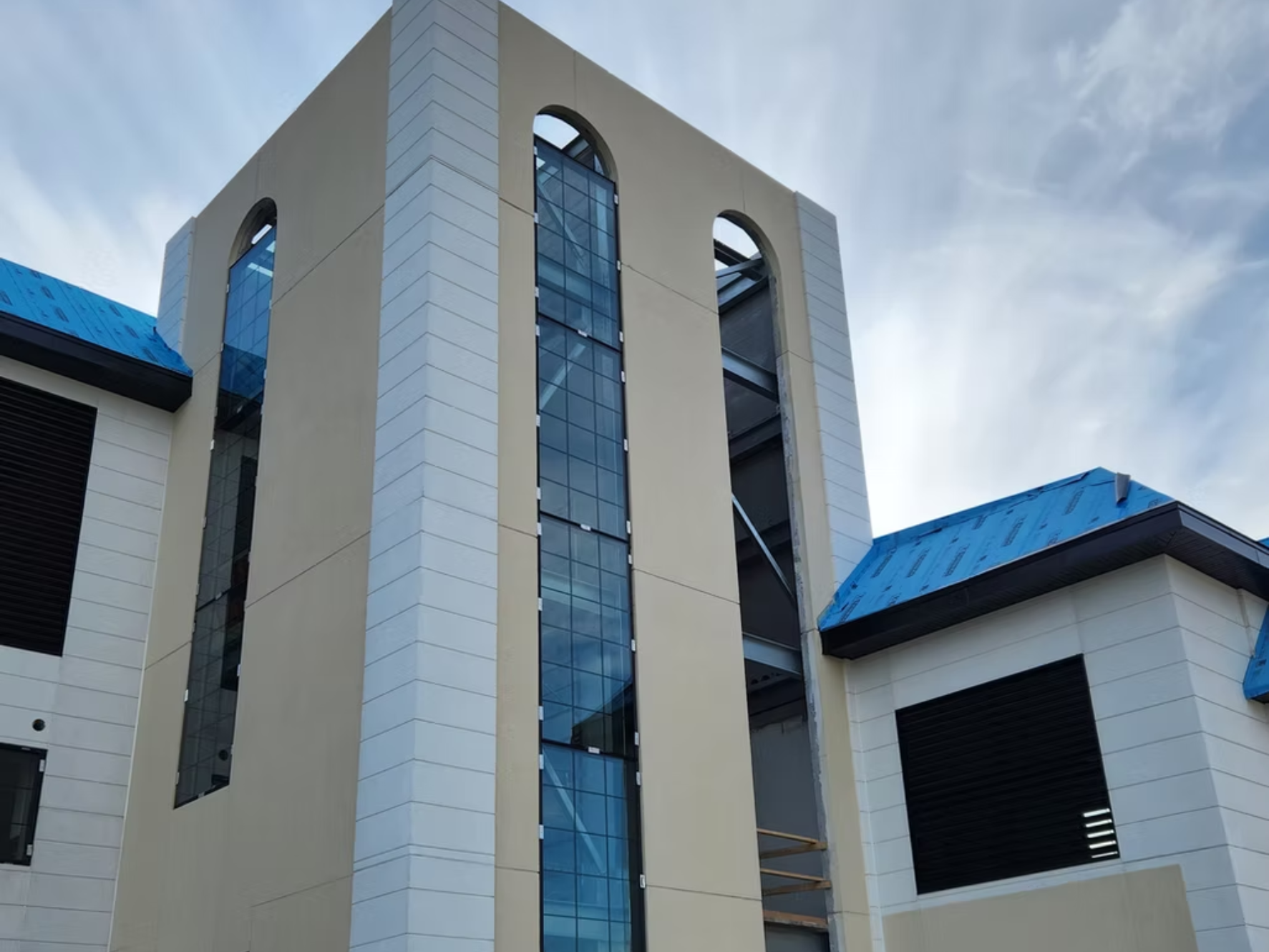 Part of a modern building with beige walls, large vertical glass windows, and a blue roof with solar reflective coating under a cloudy sky.
