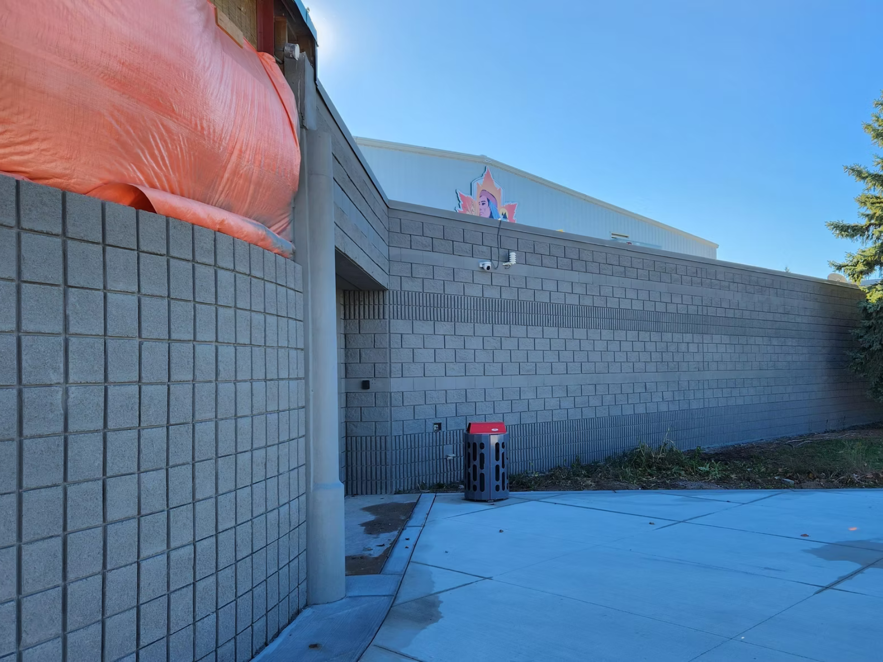Exterior view of a modern building wall made of concrete blocks with a section covered by orange tarp. A security camera and a rain sensor are mounted on the wall. There is a trash can on the sidewalk and a large sign with a woman's face and a colorful background visible at the top of the building. A tree is seen on the right side of the image, and the sky is clear and blue.