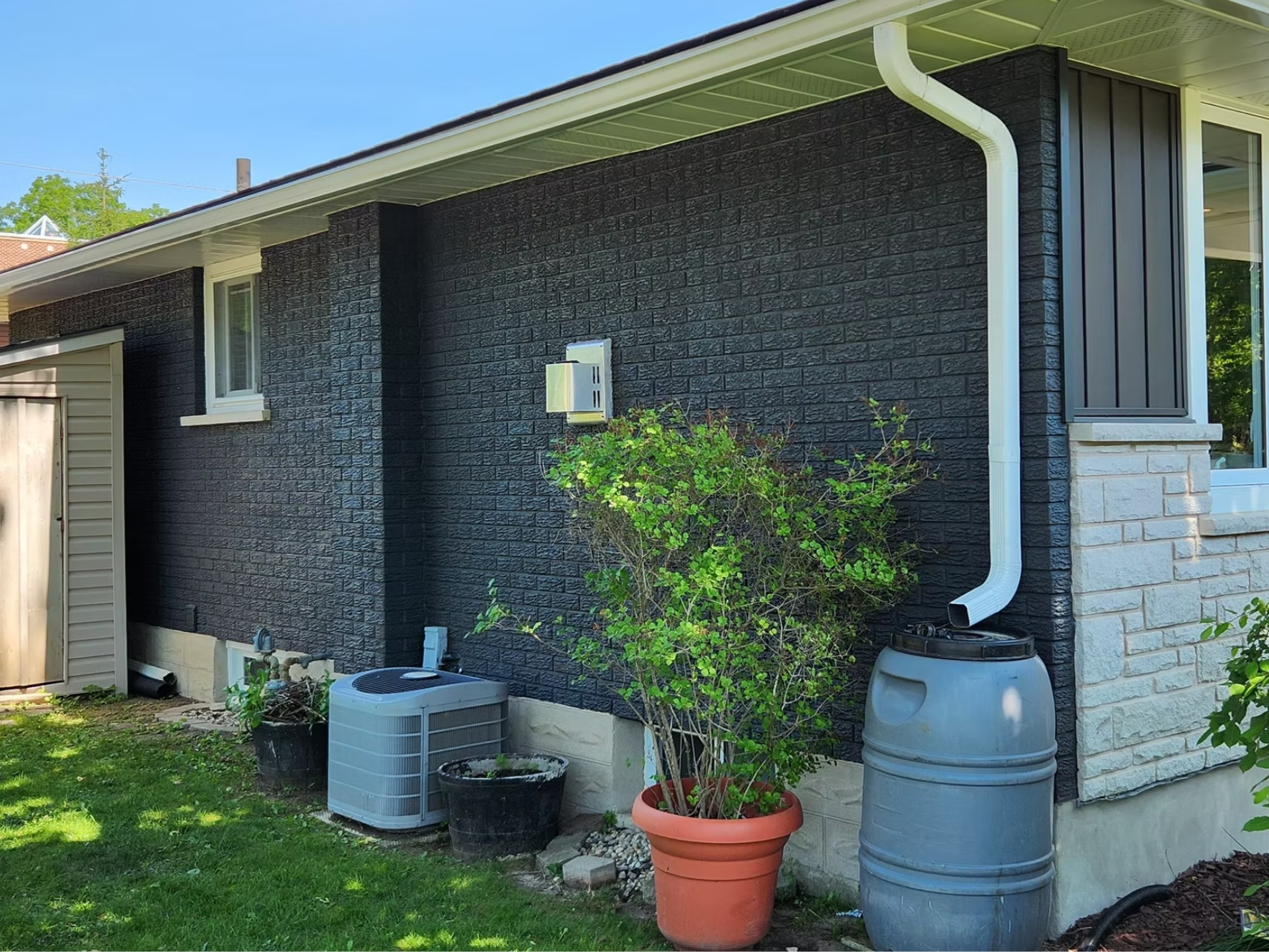 The side of a house with a black brick exterior, a small white window, white rain gutter system, and an attached white brick section. There is a gray barrel-style rain collection barrel, an air conditioning unit, and a large terracotta pot with a green bush. Several smaller black pots and gardening supplies are also visible, along with green grass and a small tree or shrub in the yard.
