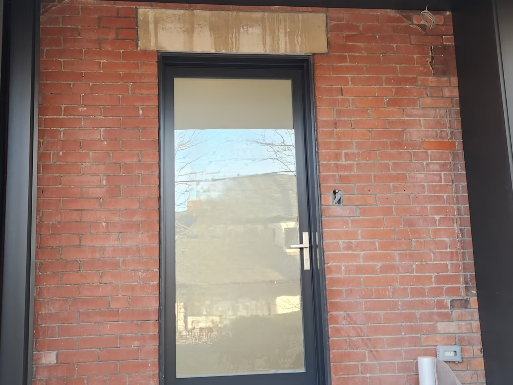 A brick wall with a black-framed glass door and a beige stone lintel on top. A power outlet and a white object are visible at the bottom right corner.