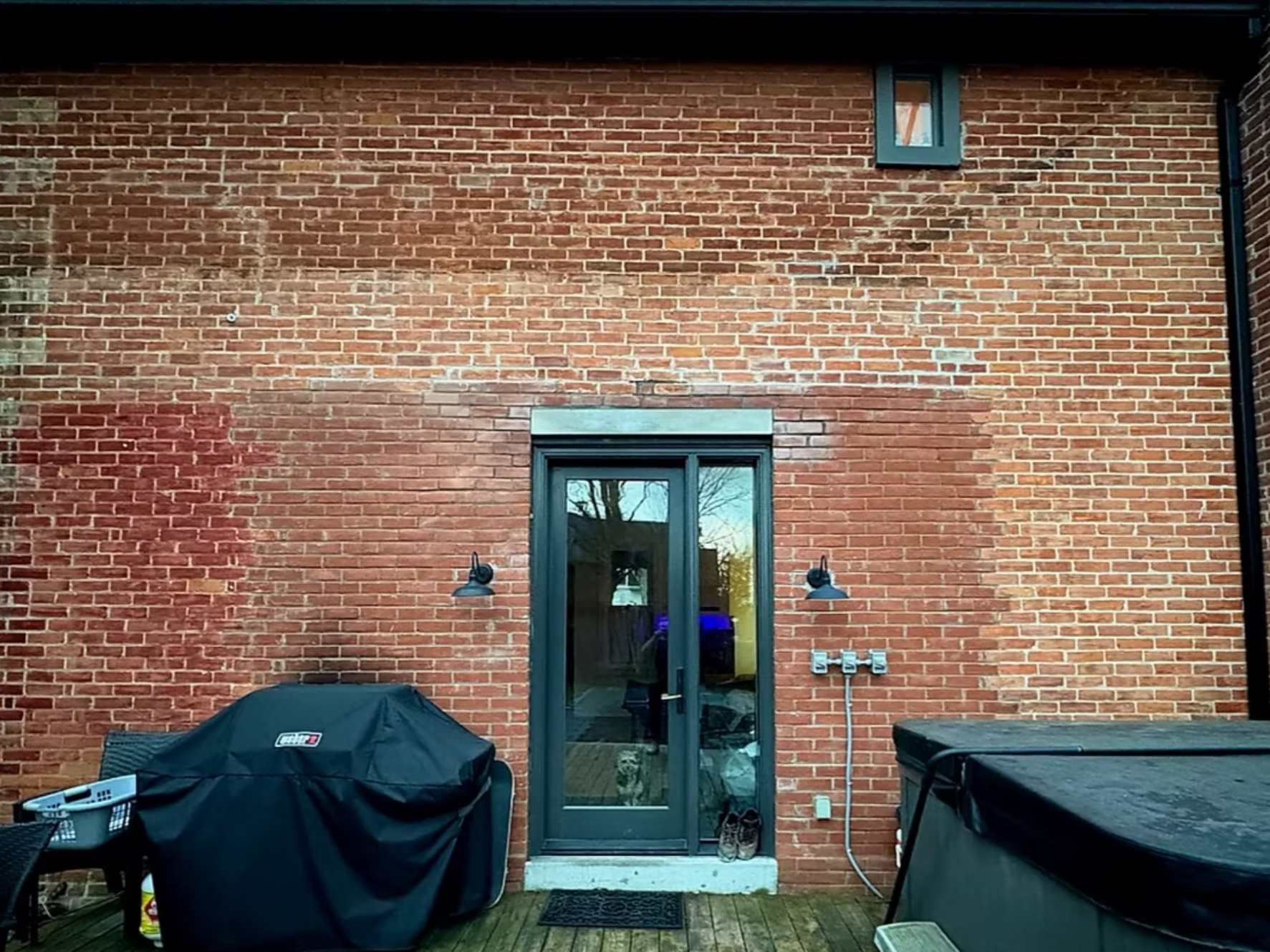 Back patio with black metal door, outdoor grilling area covered with black grill cover, and various outdoor furniture, against a red brick wall with two black outdoor lights and a small window.