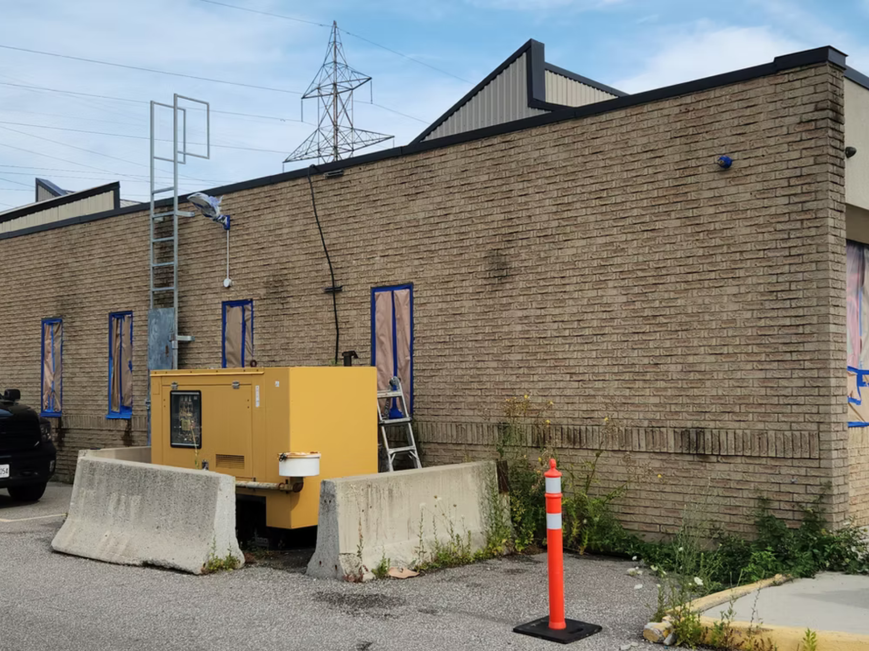 Construction site with a brick building, safety barriers, a yellow generator, a ladder, and construction cones.