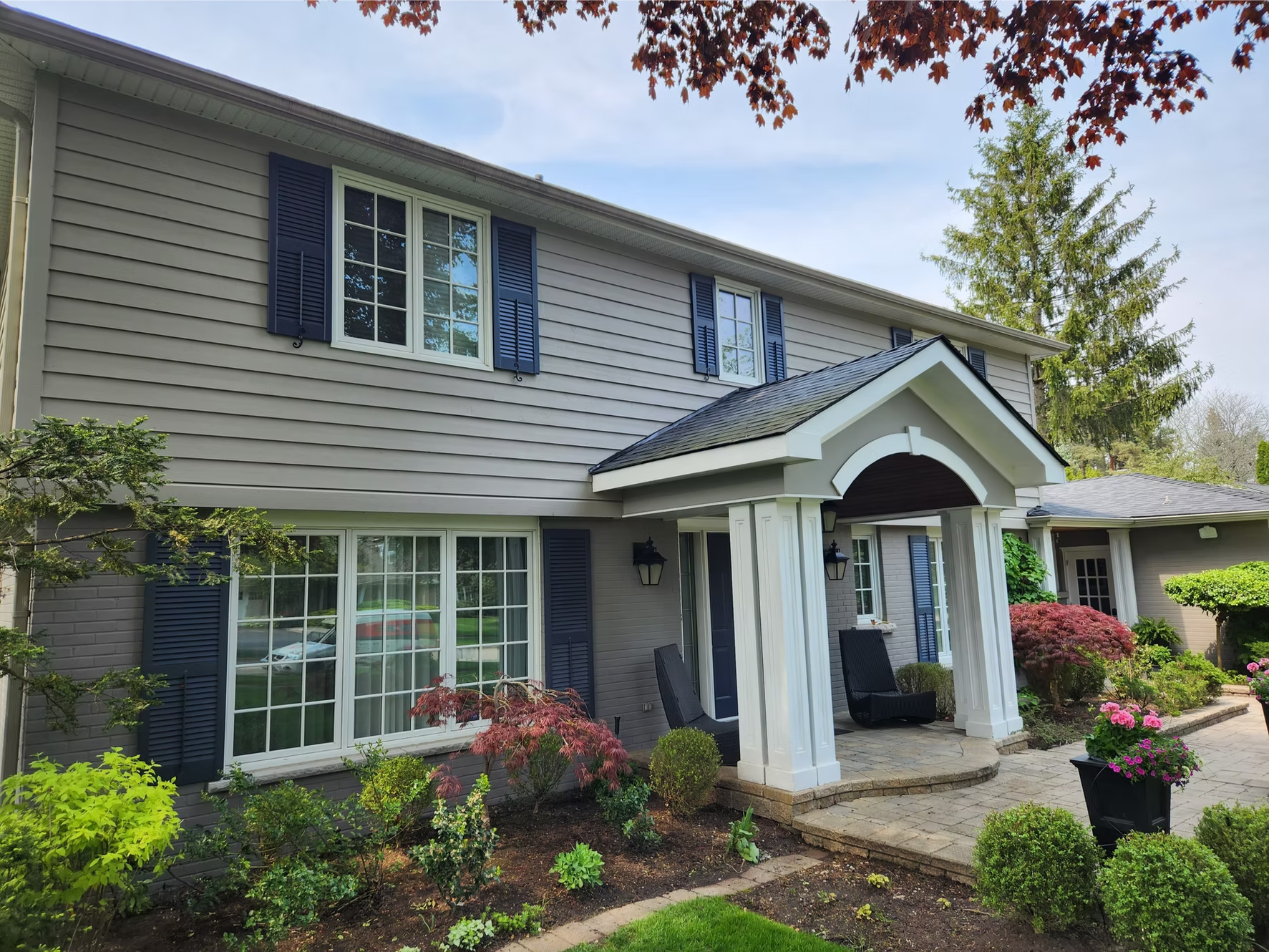 Photo of a house with gray siding, dark blue shutters, and a white portico entrance surrounded by a lush garden with various green plants, shrubs, and colorful flowers, under a partly cloudy sky.
