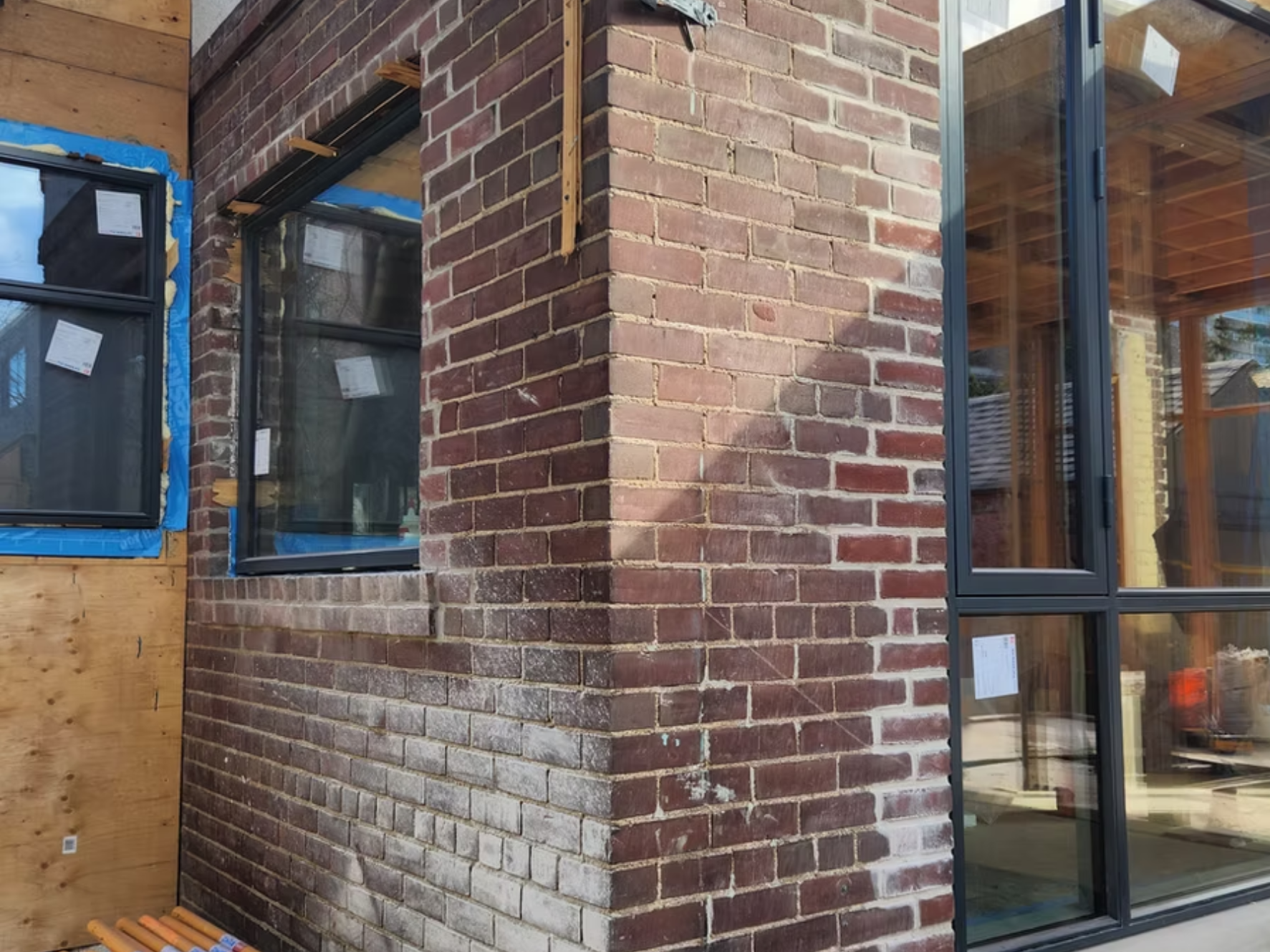 Close-up of a brick wall corner of a building under construction, with large black-framed windows and exposed wooden interior beams visible through the glass.