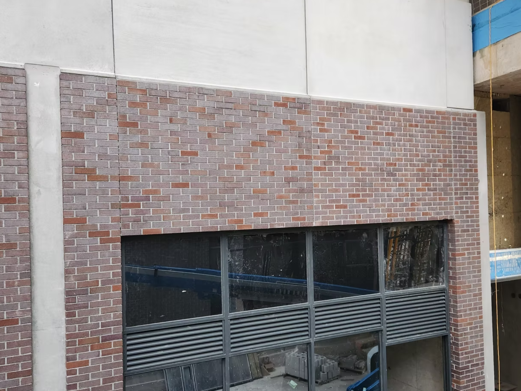 Under construction building with brick facade, large glass windows, and metal louvered vents, with construction materials visible inside.