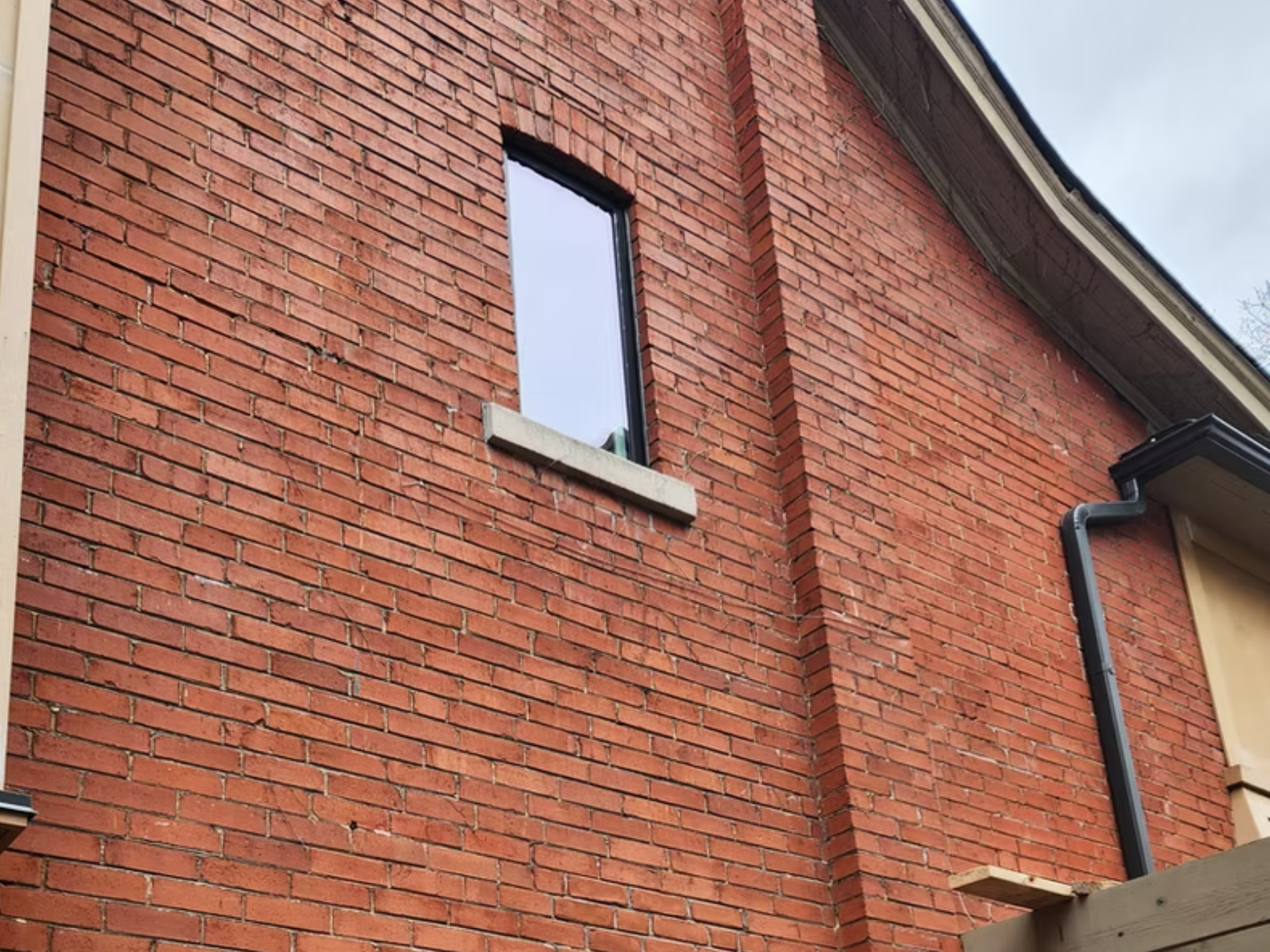 Exterior view of a red brick house wall with a small vertically rectangular window, a section of a gutter, and part of a wooden deck or staircase.