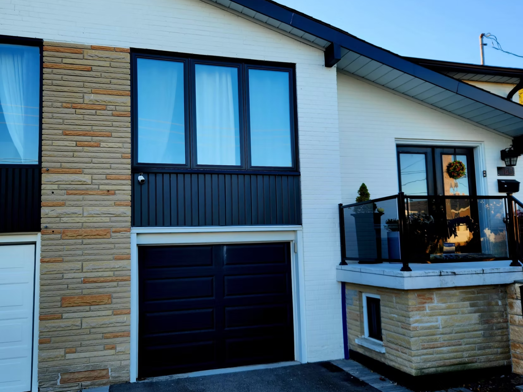 Front exterior of a modern house with a black garage door, a large window with blue curtains, a small porch with a wreath on the door, and a mix of white siding and tan brickwork. There is also a balcony with black railing and potted plants.