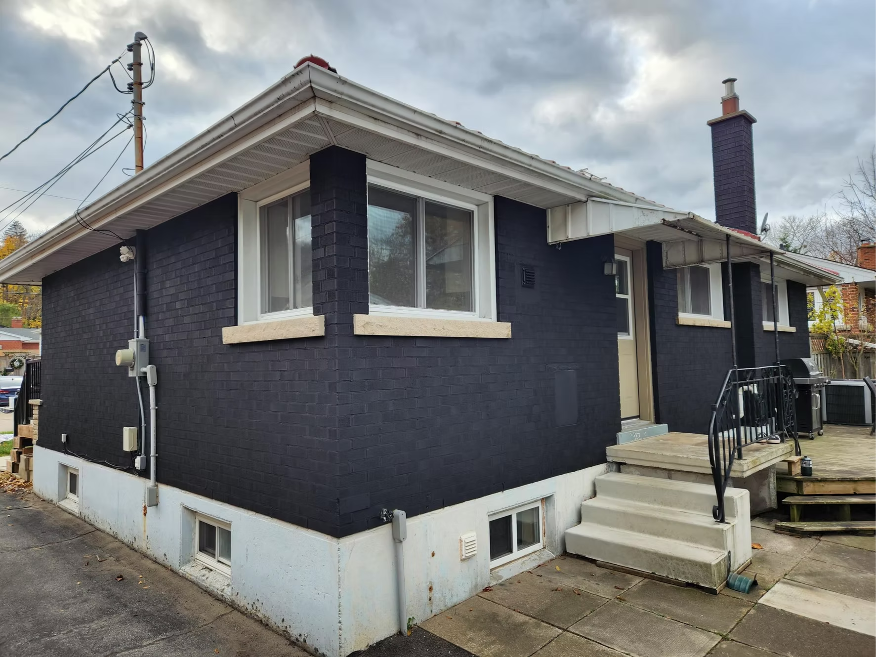 The back of a house painted black with white trim, featuring multiple windows, a chimney, and stairs leading to the door, with an overcast sky.