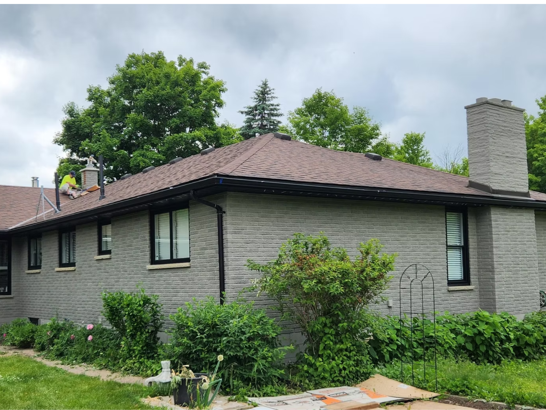Man installing roof shingles on a house roof, with trees and gray sky in the background.