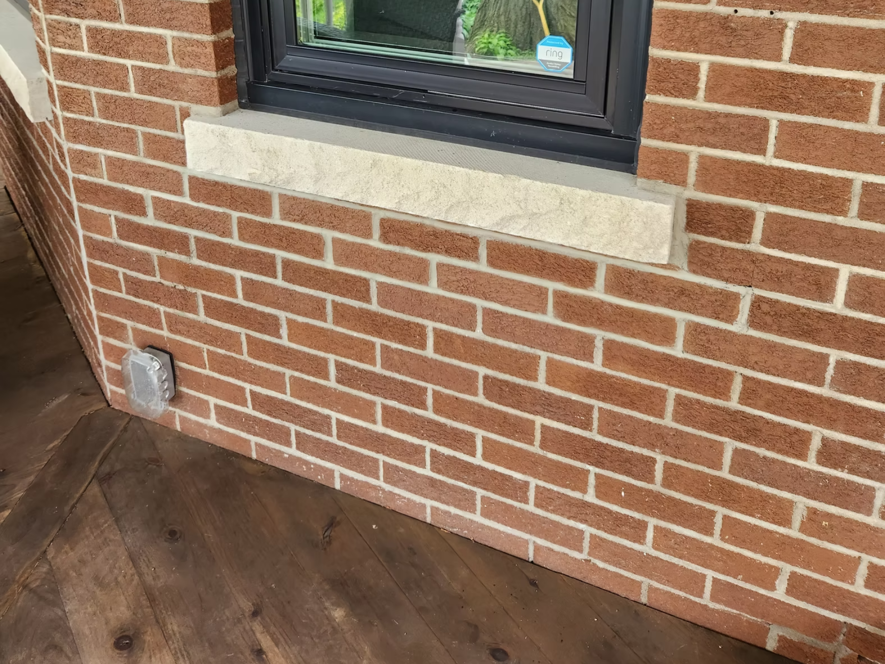 Close-up of a brick house wall with a window and a stone window sill. There is an outdoor electrical outlet near the bottom left corner of the wall and wooden deck flooring in front.