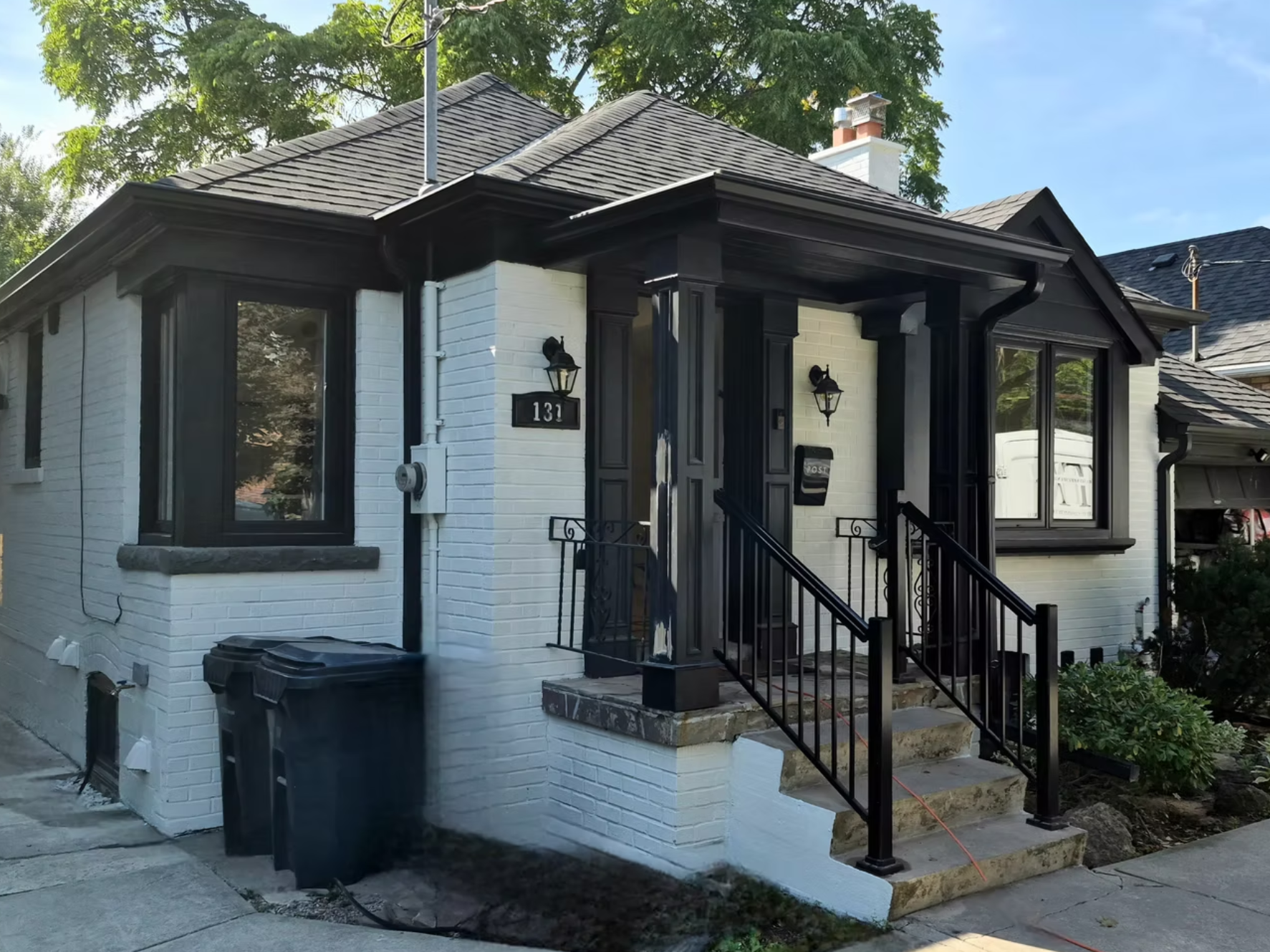 Front view of a white brick house with black trim, stairs, and outdoor lighting fixtures, in a residential neighborhood under a partly cloudy sky.