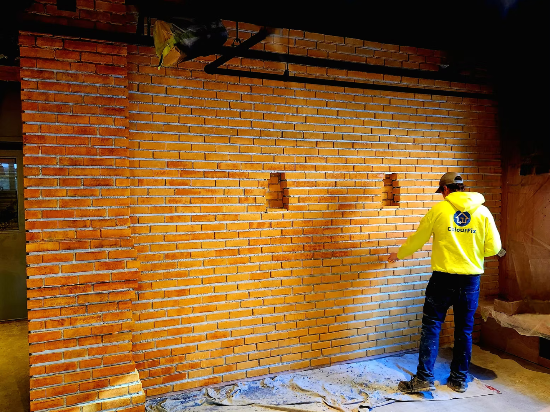 A worker in a yellow hoodie with the logo 'ColourFix' is standing in front of a brick wall under construction, with the wall featuring three small rectangular openings. The worker appears to be completing or inspecting the work. The setting suggests an indoor construction site.