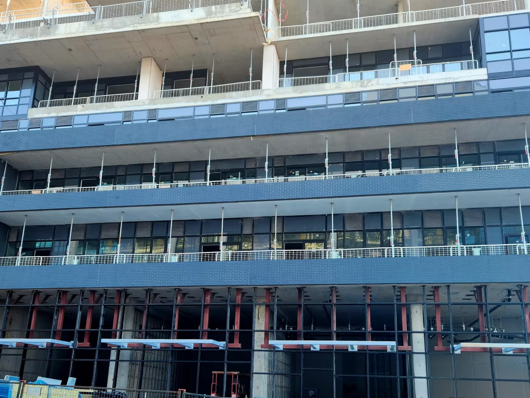 Multiple floors of a building under construction with exposed concrete, glass windows, and metal railings. Scaffolding and construction materials are visible.