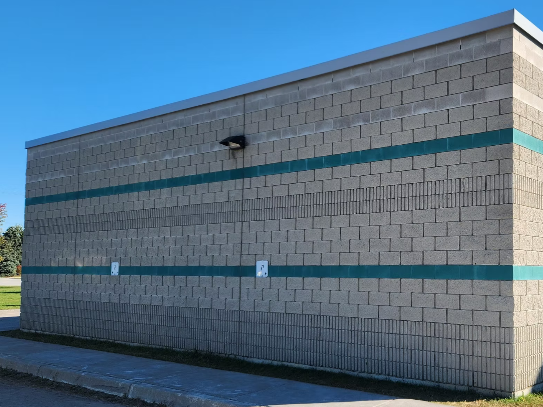 Side view of a commercial building with gray brick walls, two teal horizontal stripes, a security camera mounted on the wall, and electrical outlets installed on the lower part of the wall. There is a sidewalk and grass in the foreground with trees visible in the background.