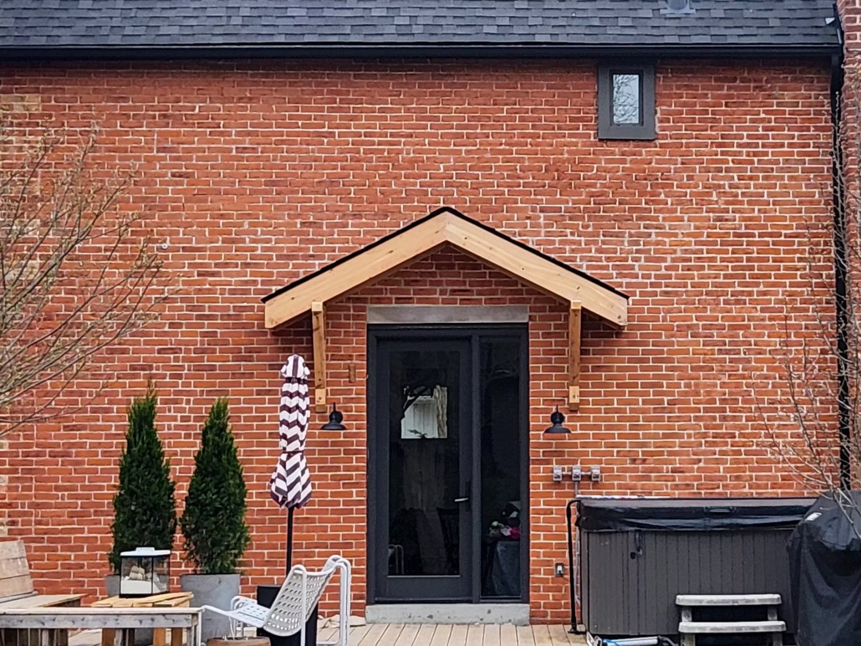 Backyard of a brick house with a black door, a small window, potted plants, a patio umbrella, outdoor furniture, a grill, and a trash bin.