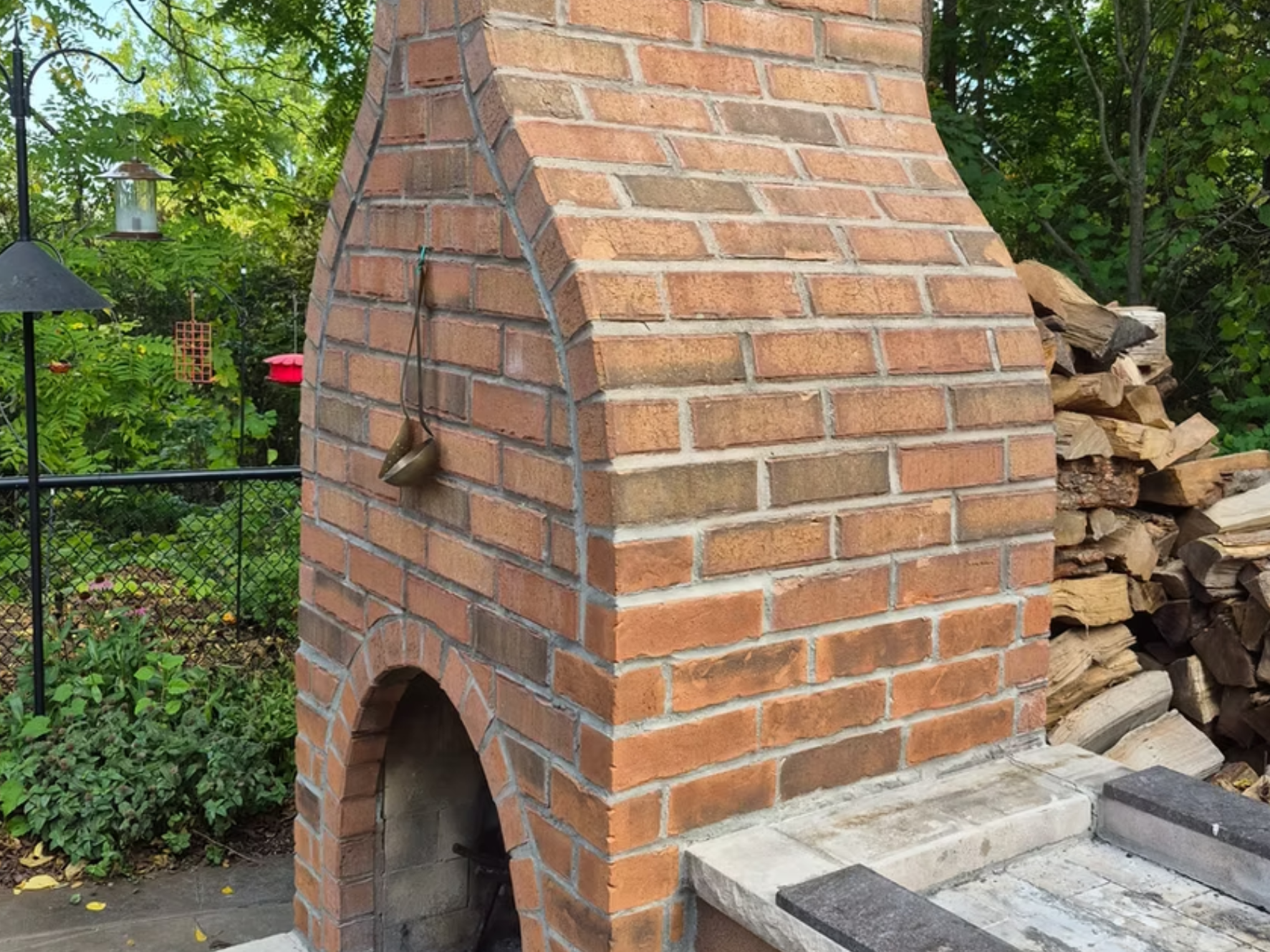 A brick outdoor fireplace with arched openings, situated on a paved patio. There is a stack of firewood beside it, and a garden area with various plants and hanging bird feeders in the background.