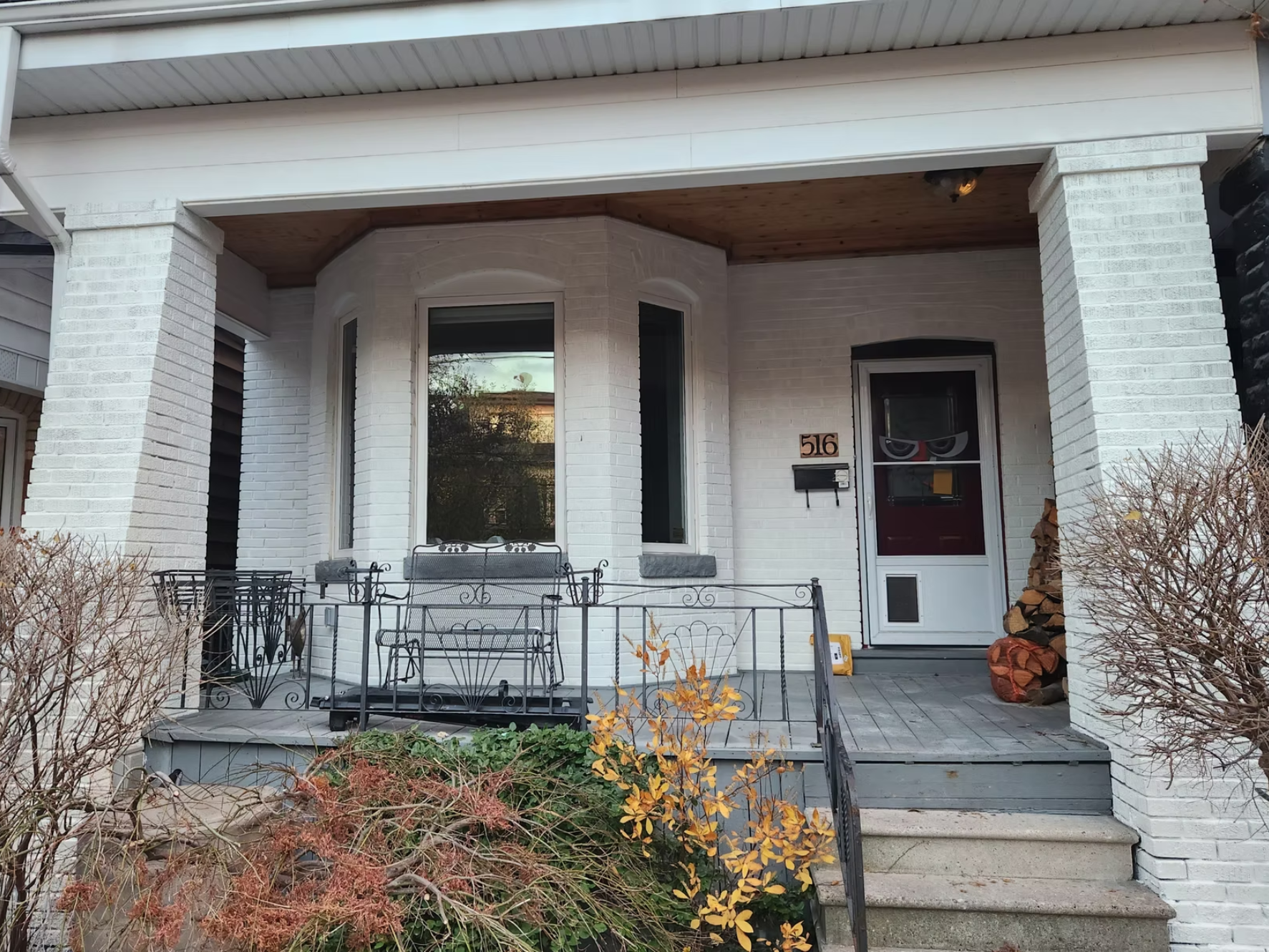 White brick house with a porch, black metal furniture, and a woodpile next to the front door. The house number is 516. There are bare bushes and plants in front of the porch.