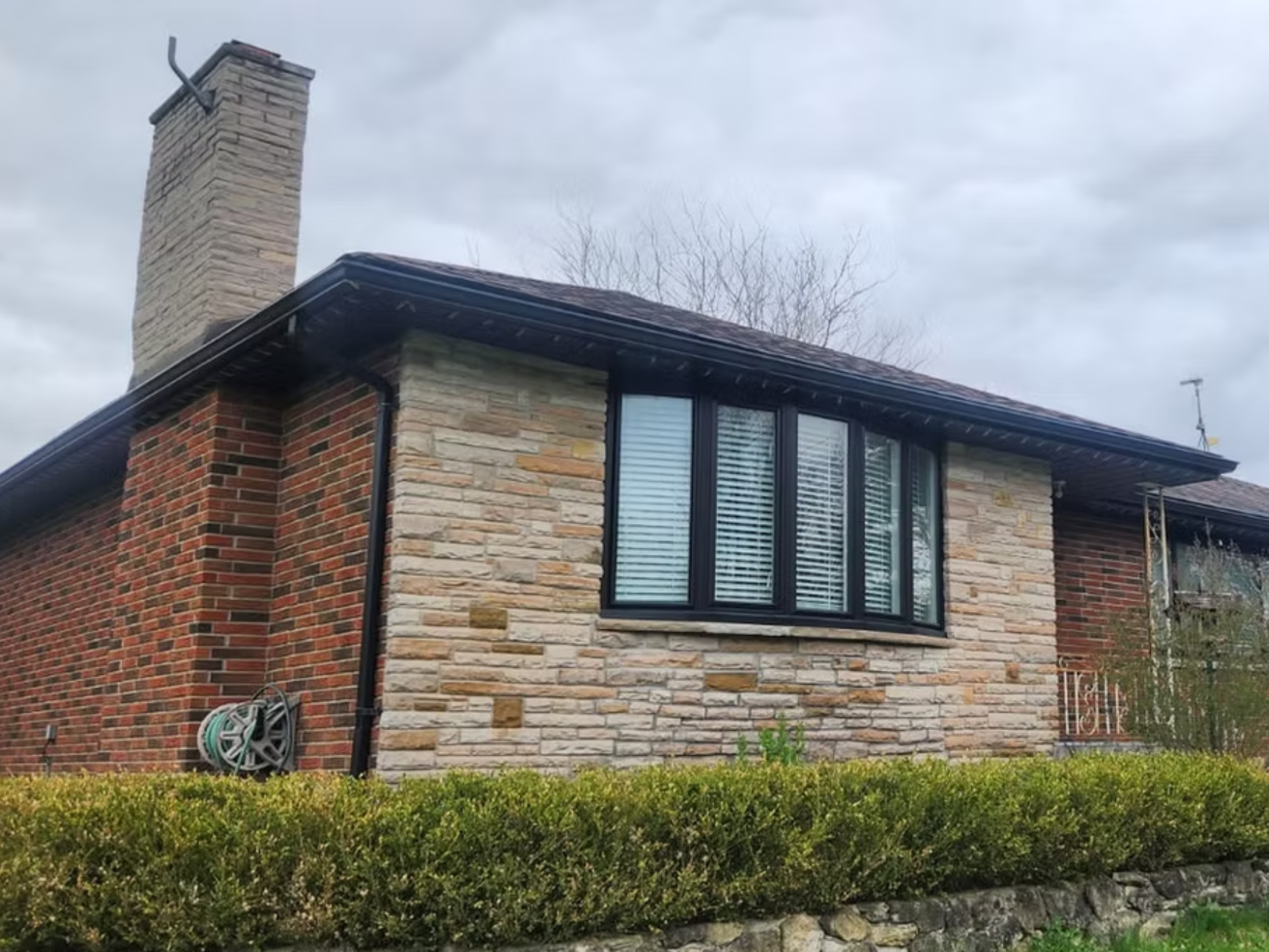 The image shows a house with a brick and stone exterior, black window frames with white blinds, a chimney made of brick and stone, and a black roof. There is a garden with green shrubs in the foreground.